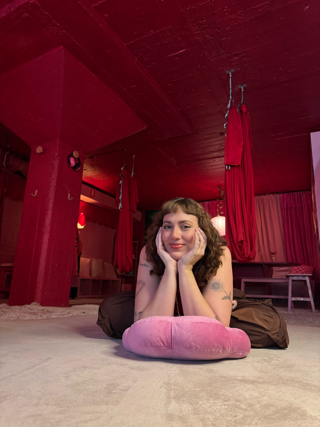A woman with curly hair, tattoos on her arms, and a slight smile lies on her stomach on a beige carpet, propping her head up with her hands. She is resting on a pink pillow in a red-themed room with curtains and hanging fabric, with a cozy and intimate ambiance.