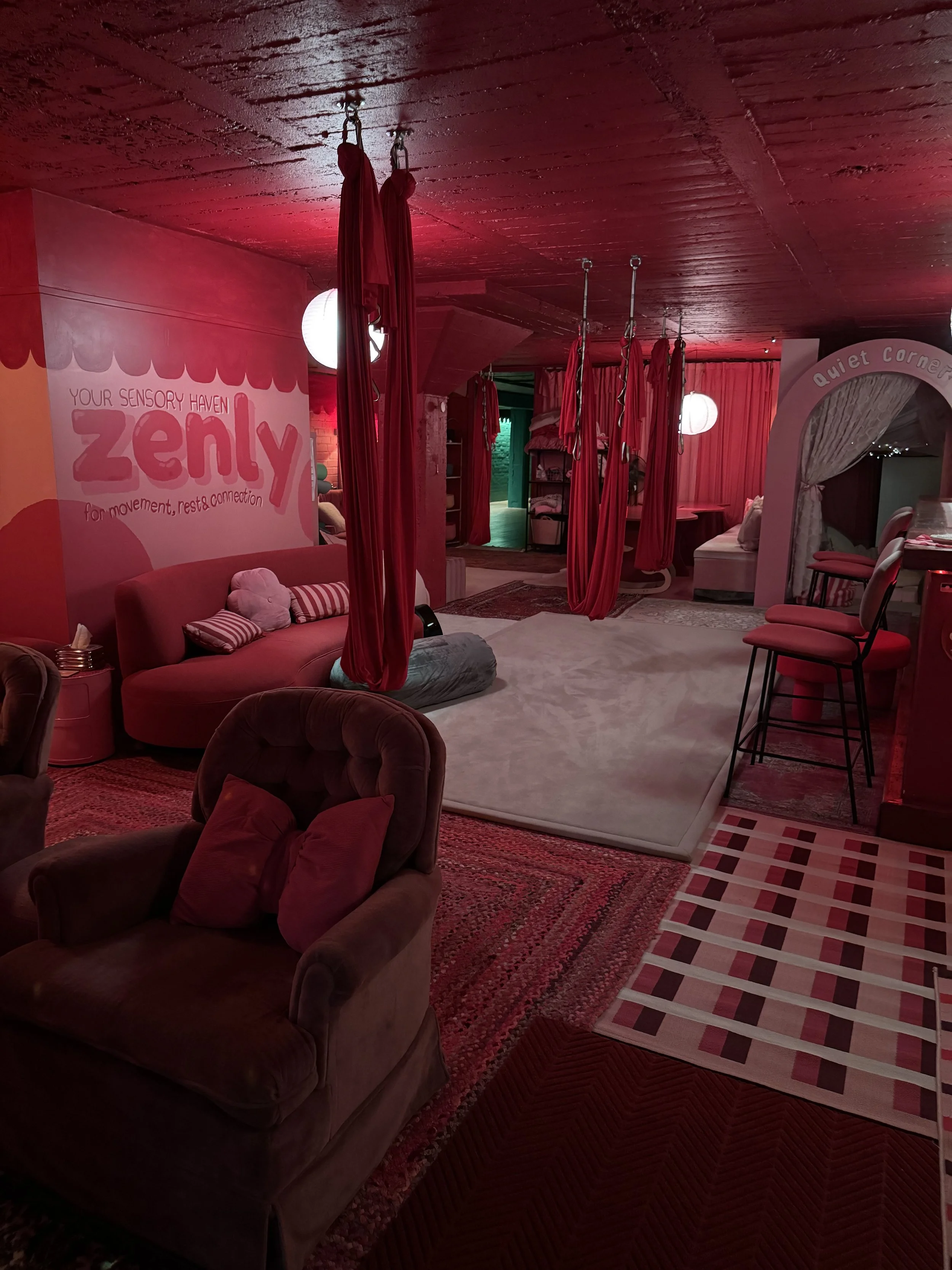 A cozy indoor space with red lighting, featuring a red couch with striped pillows, multiple hanging red fabric swings, a green-lit doorway, a bed with pillows, chairs, and various rugs and curtains.