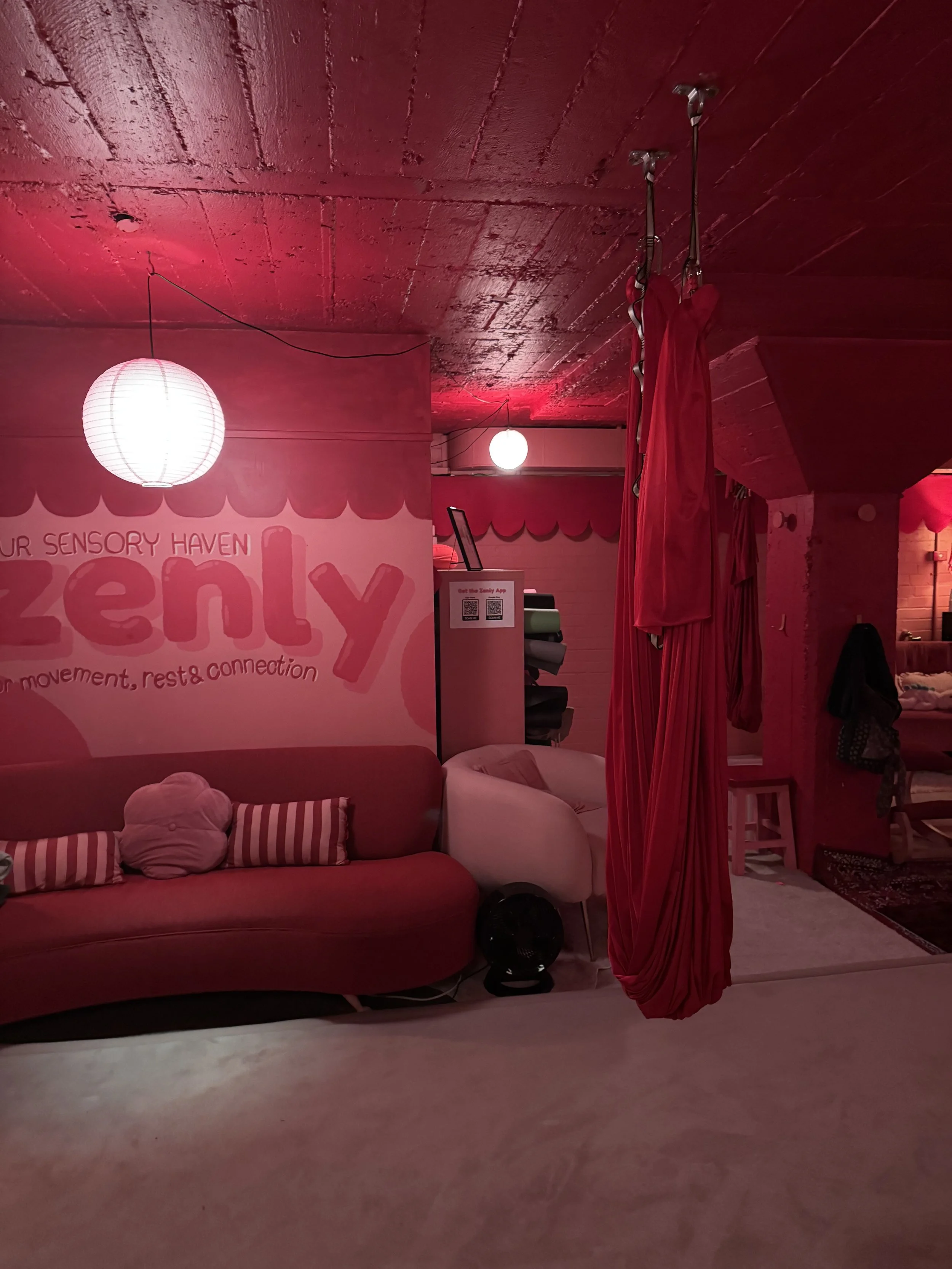 A cozy room with a red sofa, pillows, and a white chair. There are two hanging paper lanterns providing soft lighting. A red hanging fabric or hammock is suspended from the ceiling.