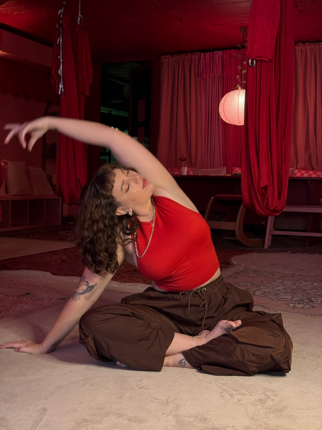 Woman with curly hair and tattoos practicing yoga in a dimly lit room with red curtains and soft lighting.