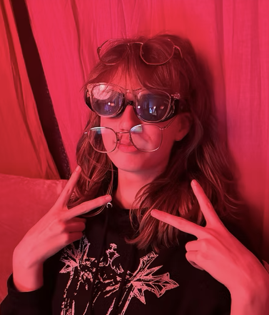 Young woman with long hair, wearing multiple glasses stacked on her face, making peace signs with both hands, in front of a red curtain backdrop.
