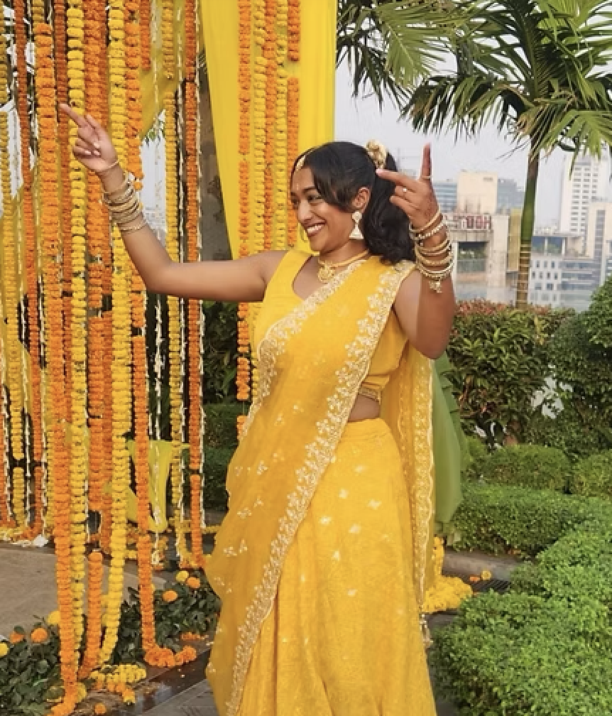 A woman in a yellow saree smiling and posing at a festive outdoor event with orange and yellow floral decorations and greenery in the background.