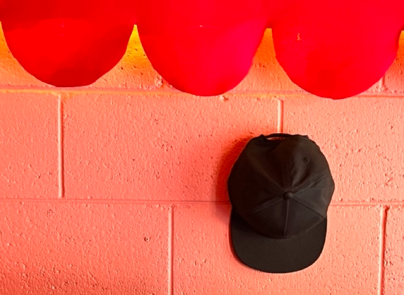 Black baseball cap hanging on a pink wall with red balloons above.