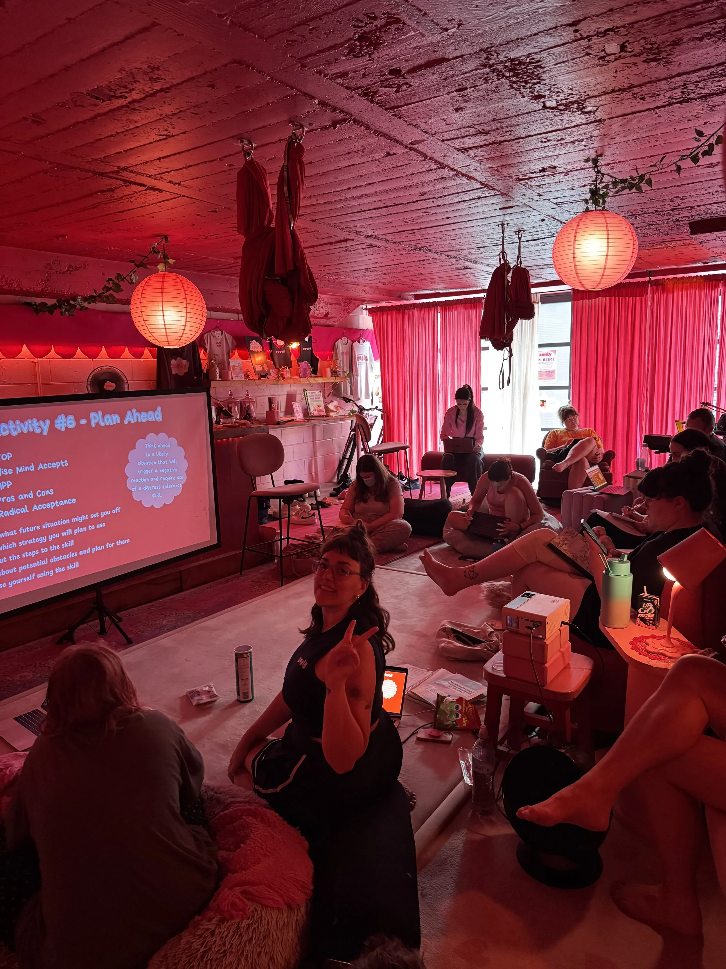 A group of people sitting on the floor and in chairs in a cozy room with red curtains and ambient lighting, engaging in a workshop or gathering, with a large screen displaying presentation slides.