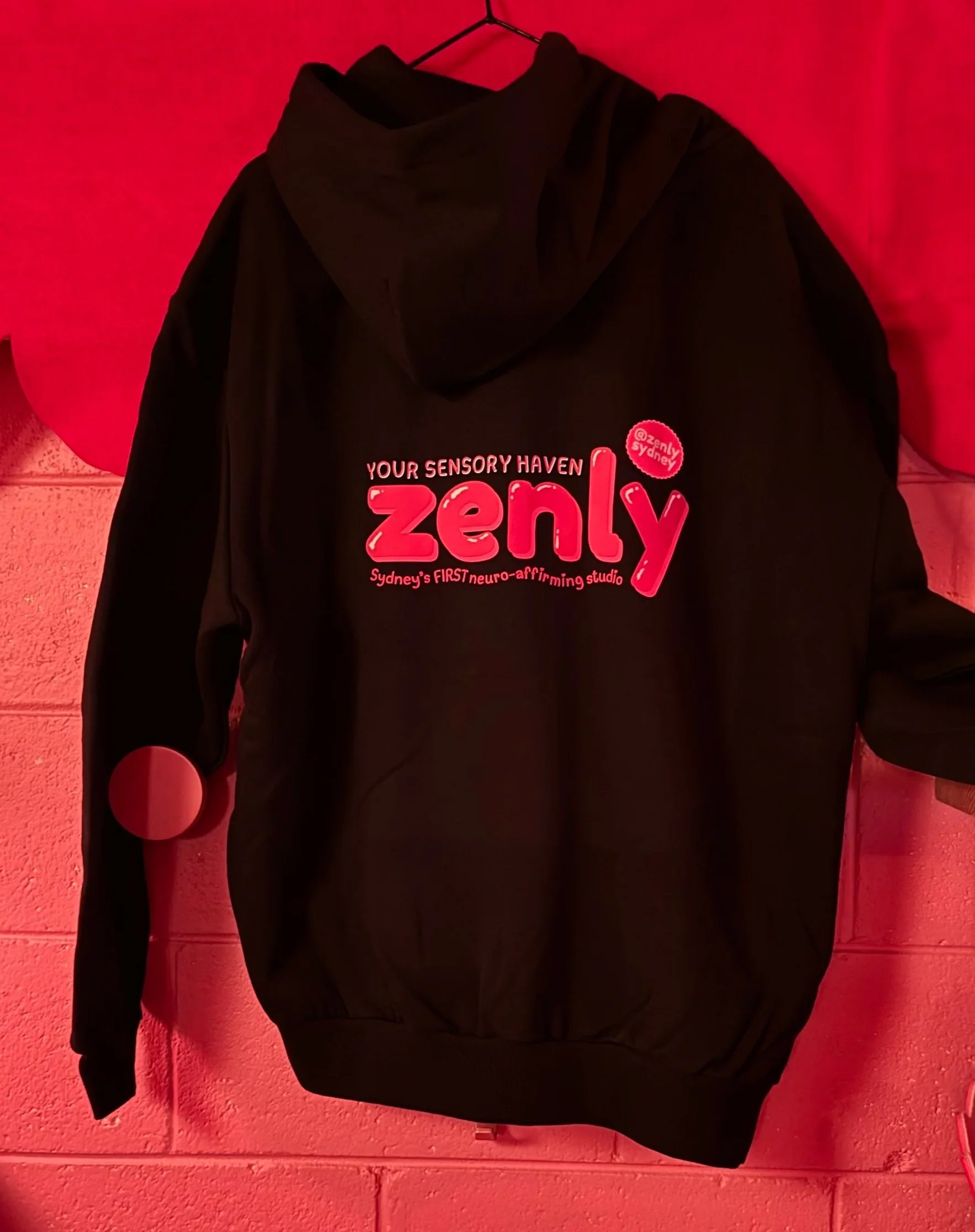 Black hoodie hanging on a hanger against a red wall with text 'Your Sensory Haven Zenly Sydney's first neuro-affirming studio' printed on it.