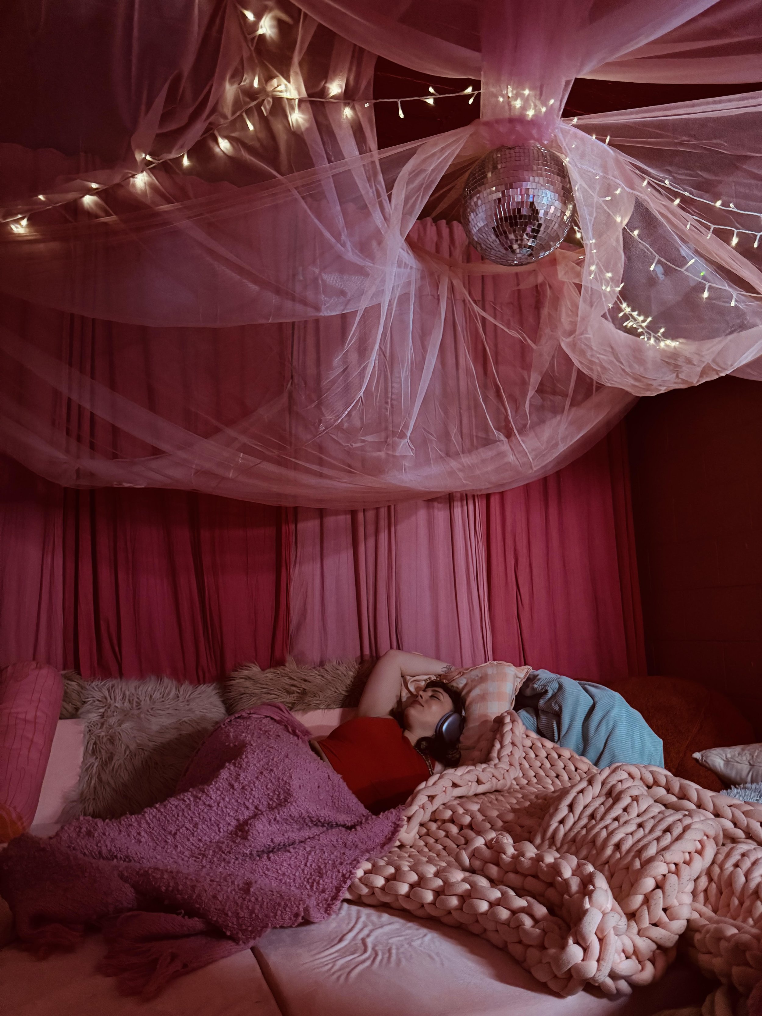 A woman is lying on a bed with pink and tan pillows, covered in a chunky pink blanket, wearing headphones, in a room decorated with pink sheer fabric, fairy lights, and a disco ball hanging from the ceiling.
