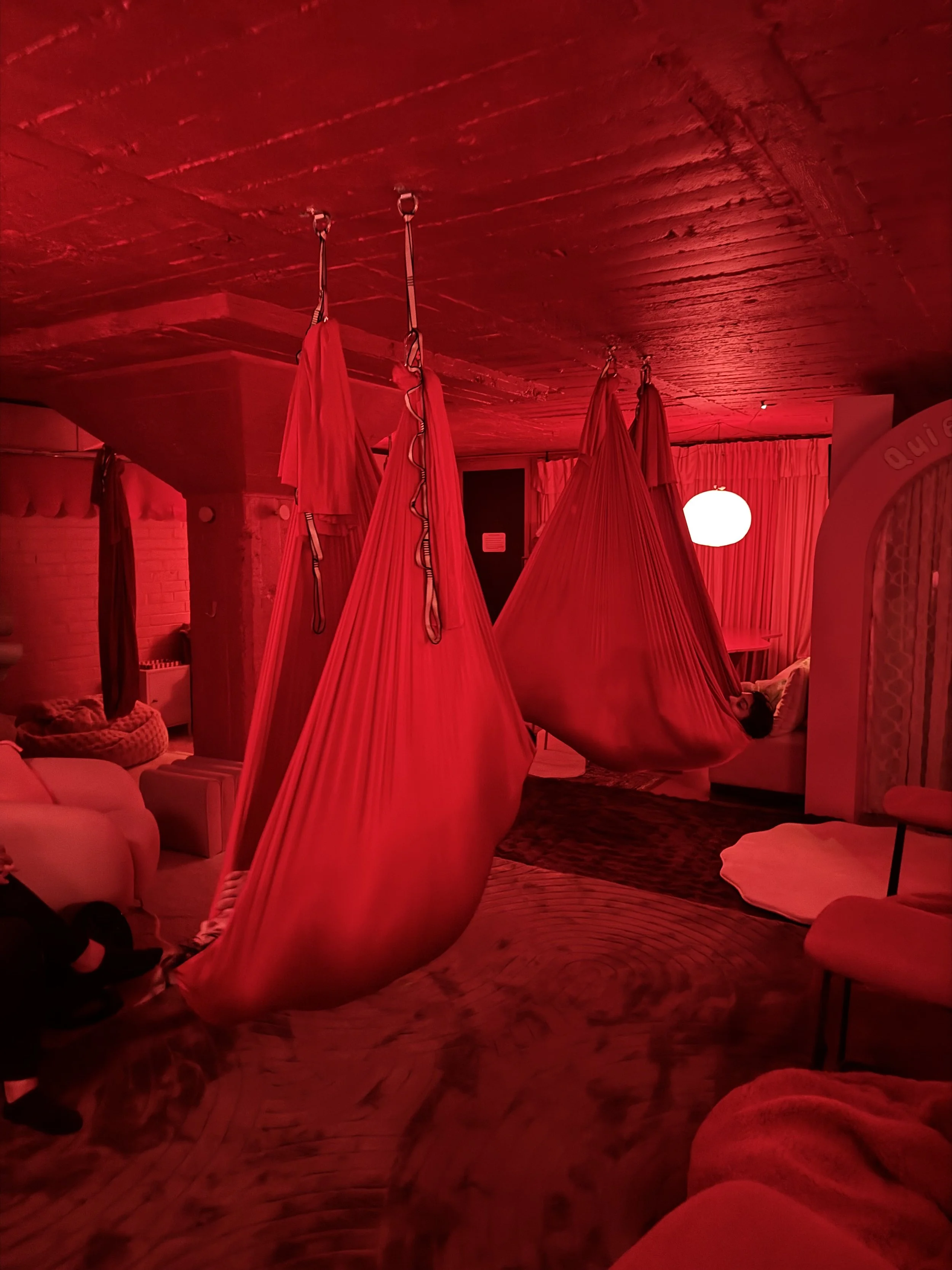 A dimly lit room with red lighting, featuring hanging fabric swings, plush furniture, a curtain-covered window, and a person lying on a swing.