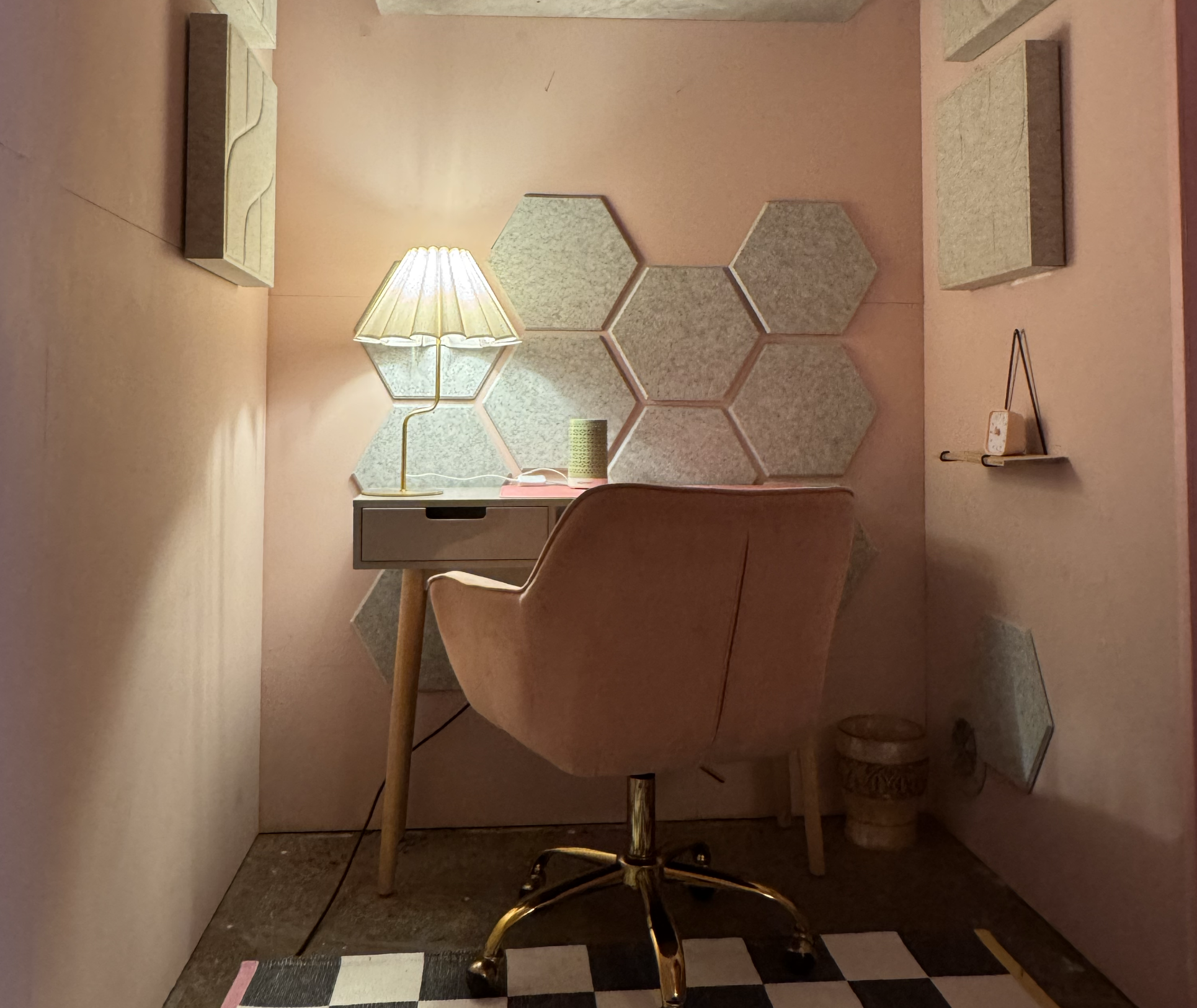 A small workspace cubicle with peach-colored walls, a beige chair, a white cabinet, a yellow table lamp, and hexagonal wall mirrors. The floor has a black and white checkerboard pattern, and there's a small trash bin under the desk.