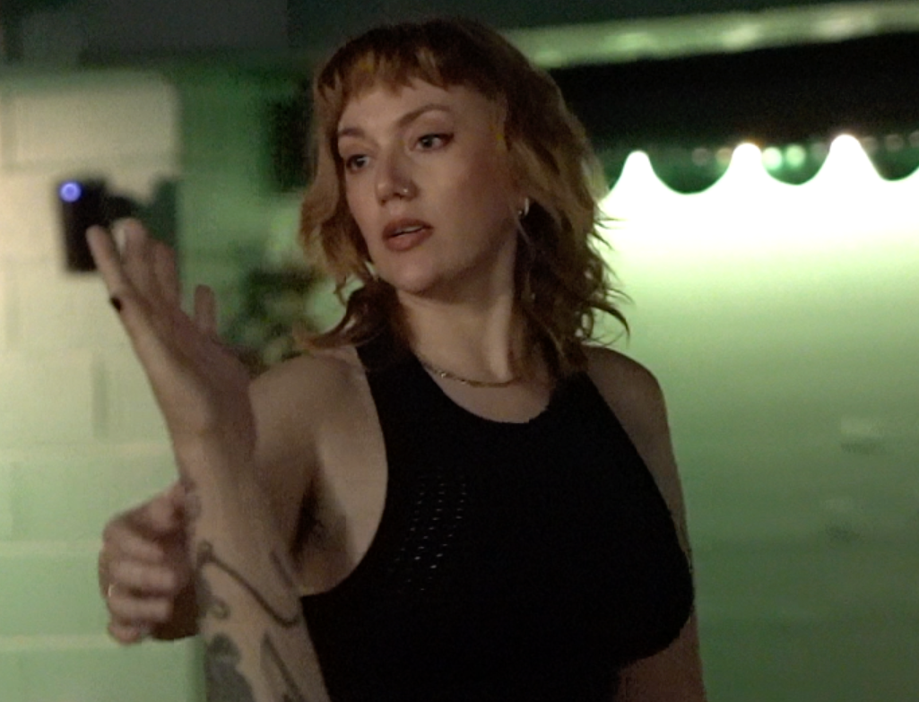 A woman with red hair wearing a black sleeveless top is holding a smartphone in her right hand, appearing to take a selfie or record herself. The background is blurry with green and yellow lighting.