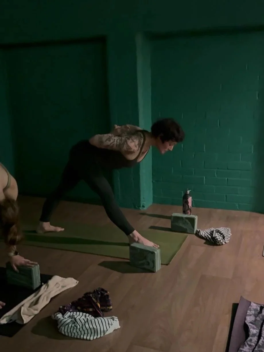 Woman practicing yoga on a green mat in a dimly lit room with green brick walls, using blocks and a towel nearby.