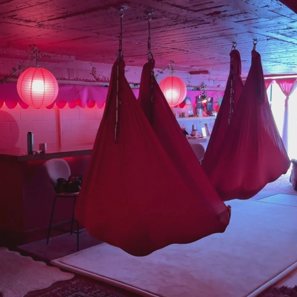 Indoor space with pink and purple lighting, featuring three hanging red fabric swings or pods, pink paper lanterns on the ceiling, a white chair with a dark cushion, a bar counter, and a shelf with photos and decorations in the background.