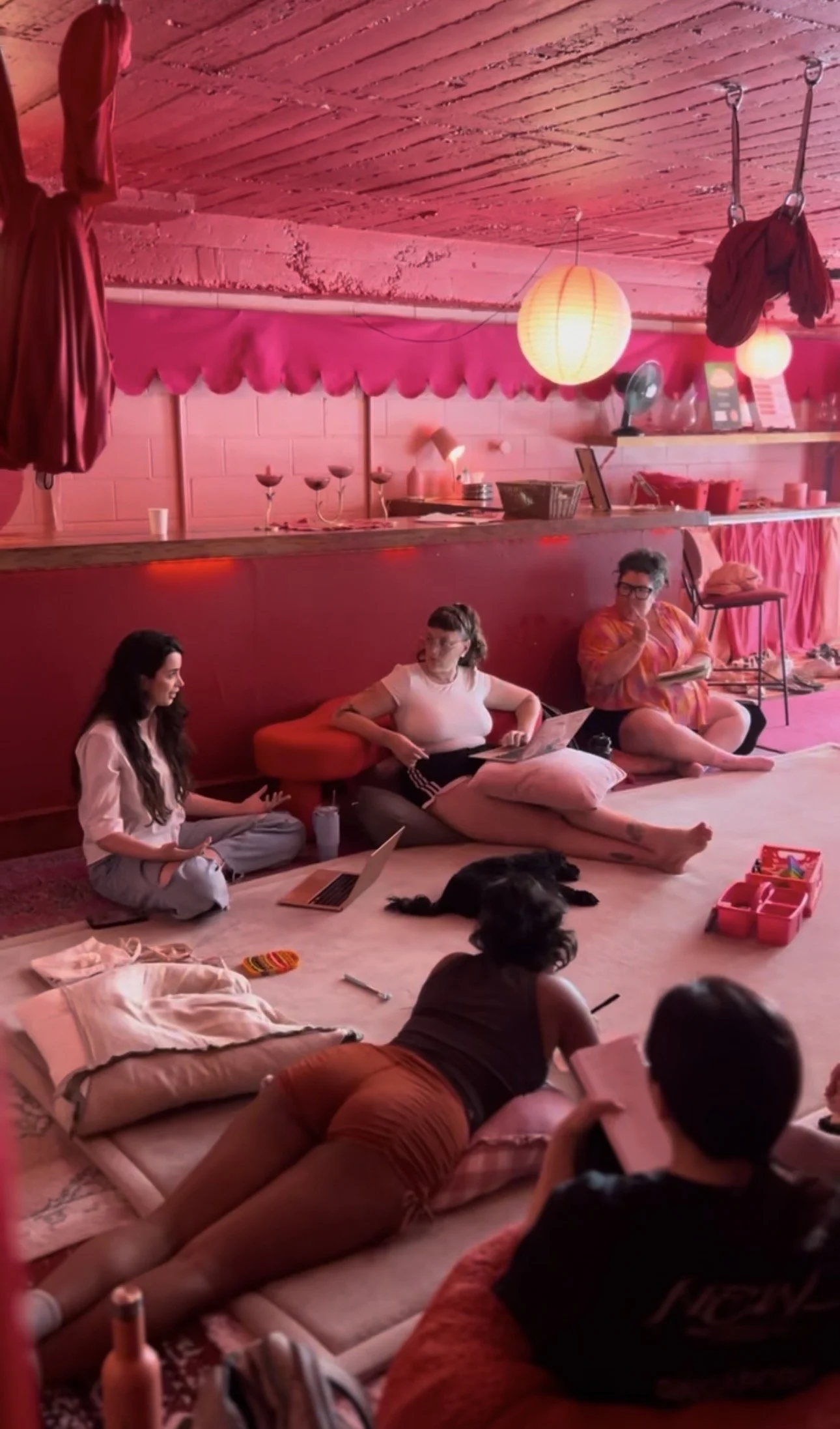 People sitting and lying on the floor in a cozy, pink-lit room with a rustic wooden ceiling, engaged in conversation or working on laptops, with various items like pillows, laptops, and art supplies scattered around.