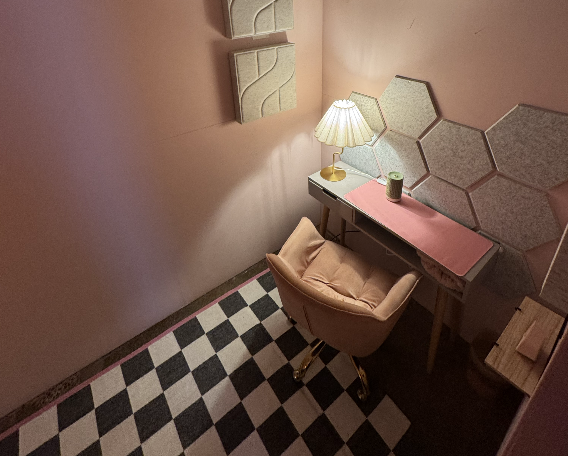 A pink wall with decorative hexagonal mirrors, a vintage-style table lamp with a pleated lampshade, a small green speaker, a desk with a pink placemat, and a tan armchair, with a black and white checkered floor.