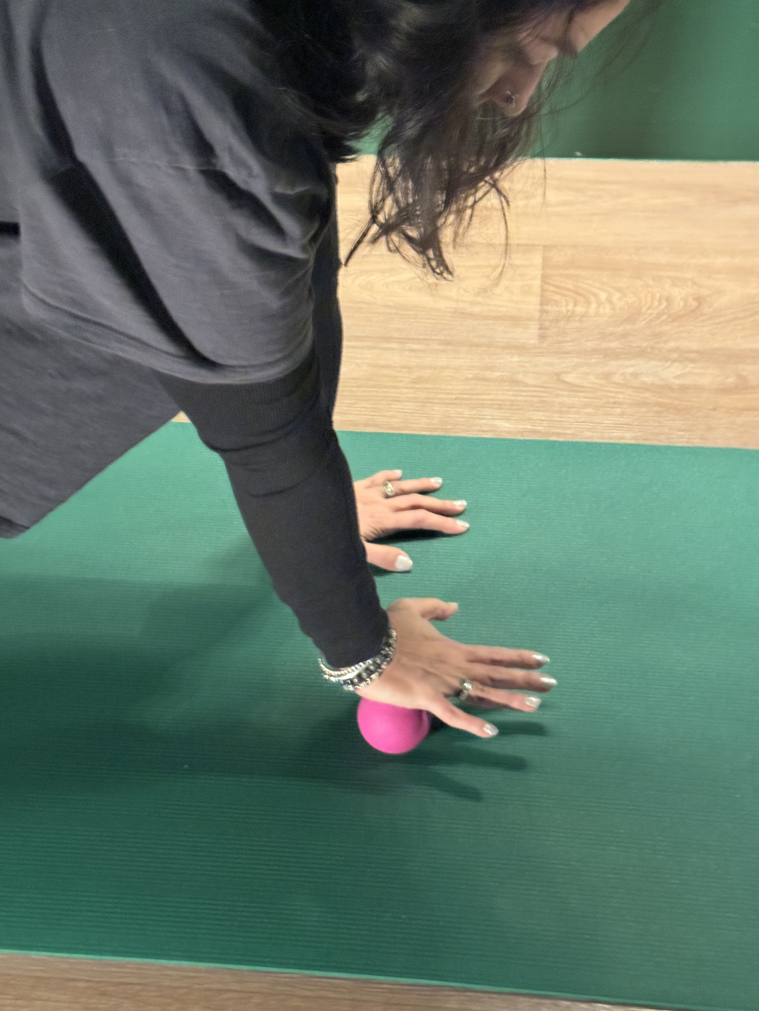 Person in black clothing doing downward dog yoga pose on a green yoga mat with a pink yoga block under their hand.
