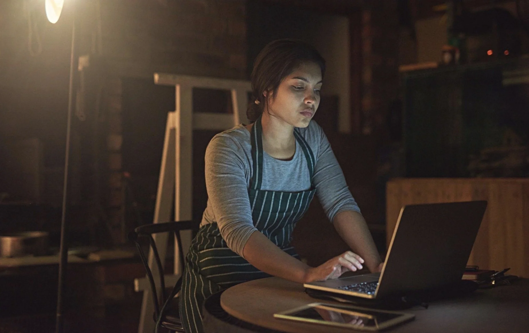 A woman in a gray long-sleeve shirt and striped apron working on a laptop at a table in a dimly lit room.