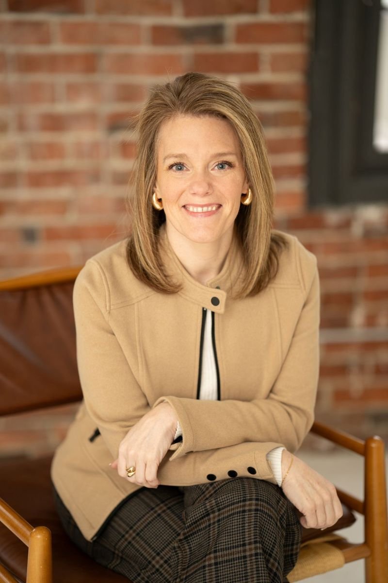 Professional portrait of Jennifer Weinmann, LMFT, a relational and couples therapist in Washington State, smiling while sitting in a chair against a brick wall.