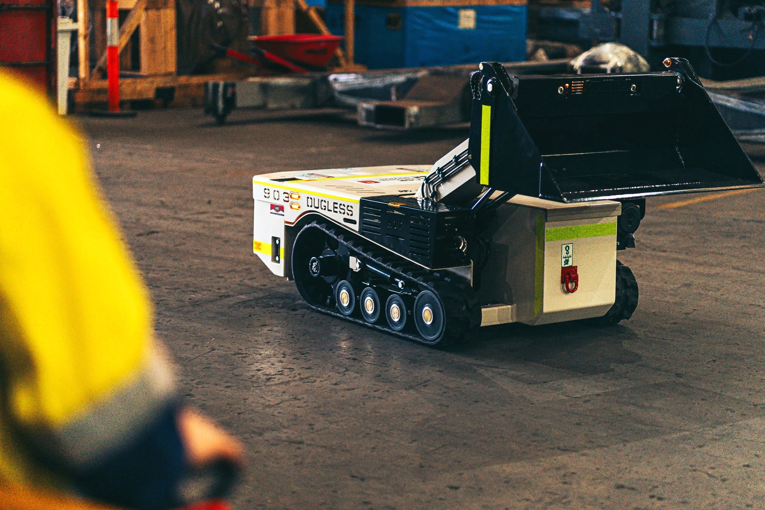 A remote-controlled toy bulldozer with tracks, labeled 'Dugless,' on a workshop floor, with a blurred yellow object in the foreground and workshop equipment in the background.