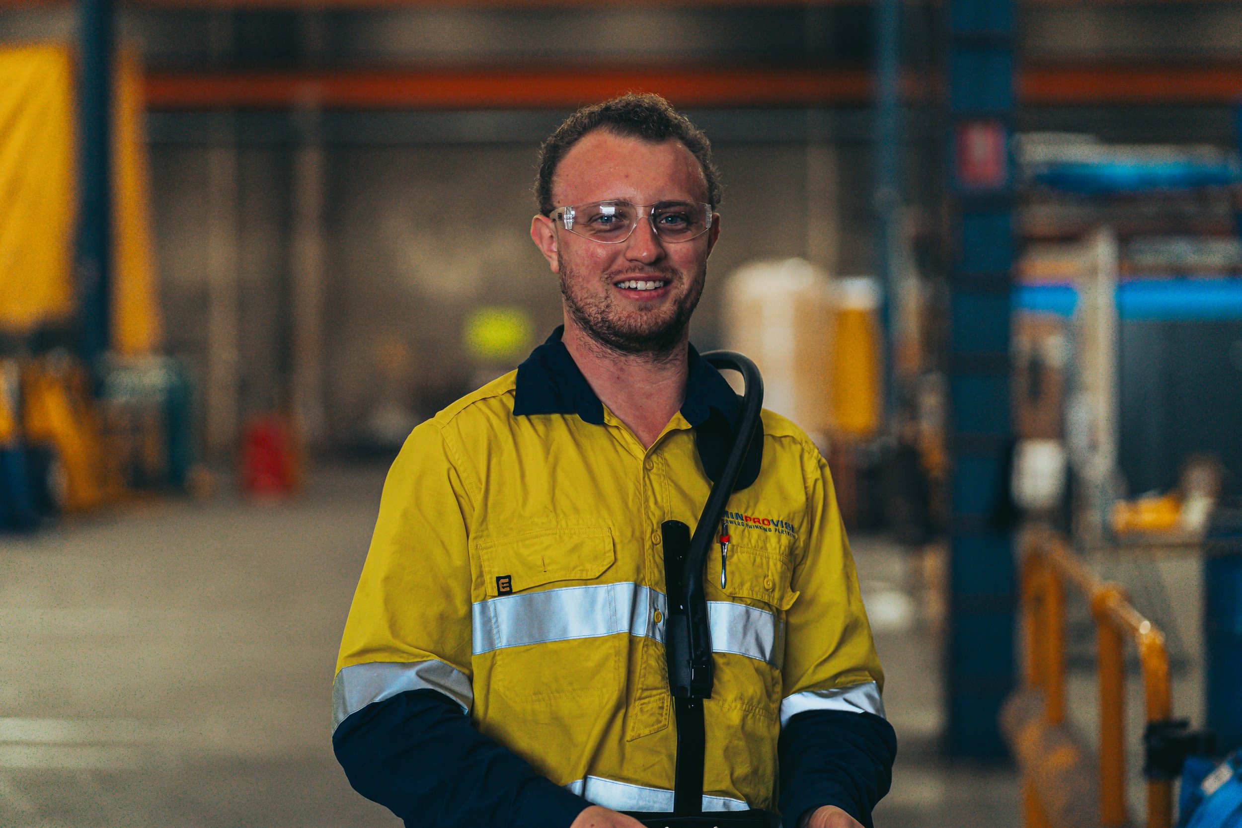 A man in a yellow safety jacket and safety glasses standing inside a warehouse, smiling at the camera.