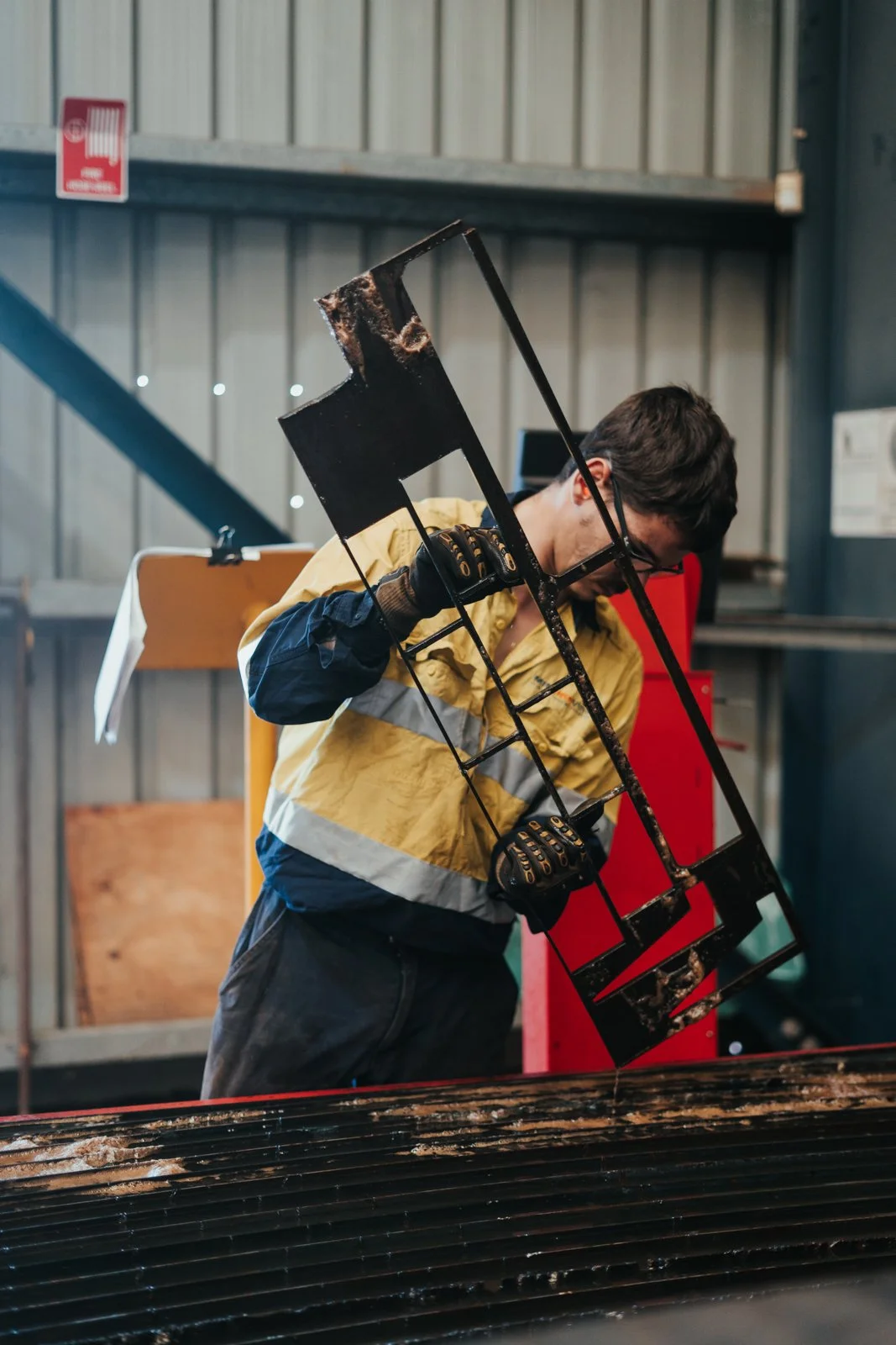 Worker in safety jacket and gloves holding a burnt metal frame, inspecting it at a workshop with industrial background.