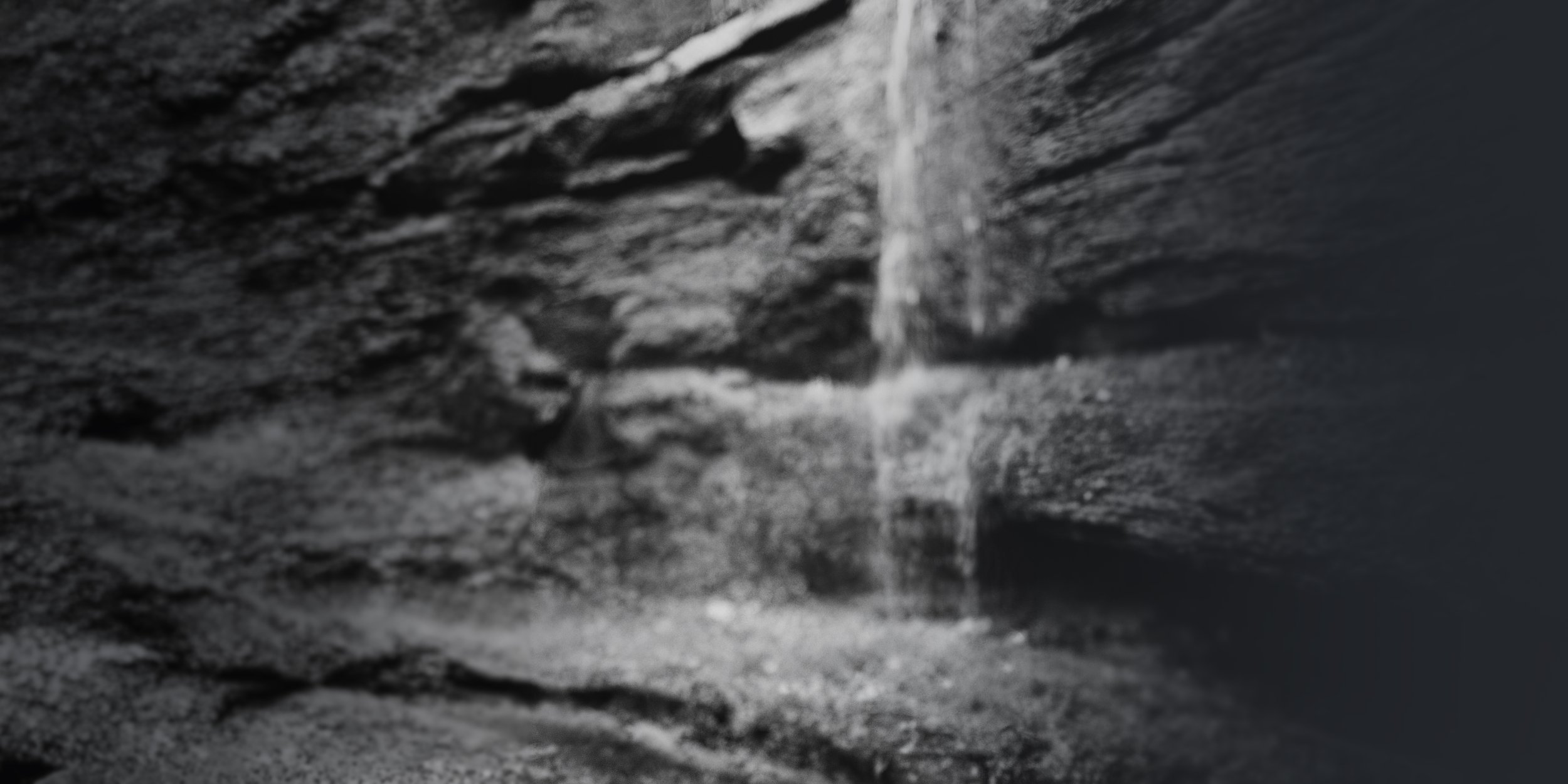 Black and white photo of a rocky cliff with a thin waterfall flowing down into a small pool of water.