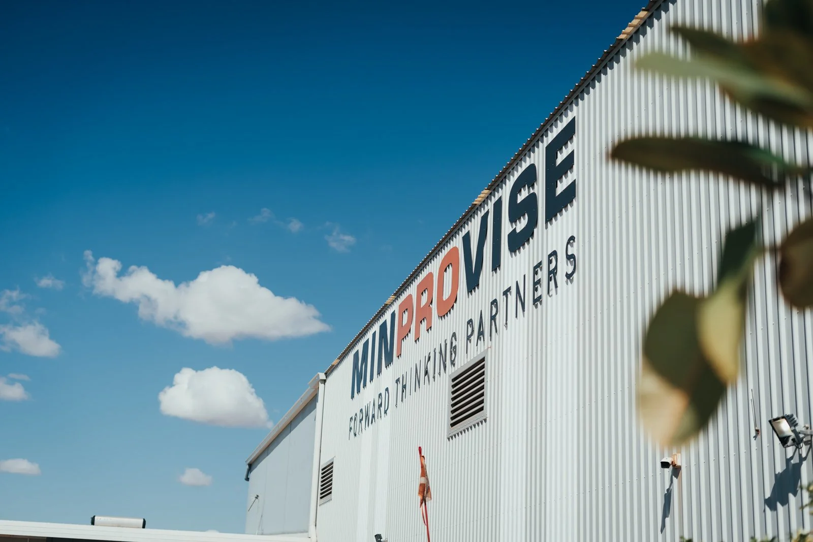 Exterior of a warehouse with the sign 'MIN PROVISO' and the words 'Forward Thinking Partners' under it, a partly cloudy sky in the background, and some leaves on the right side.