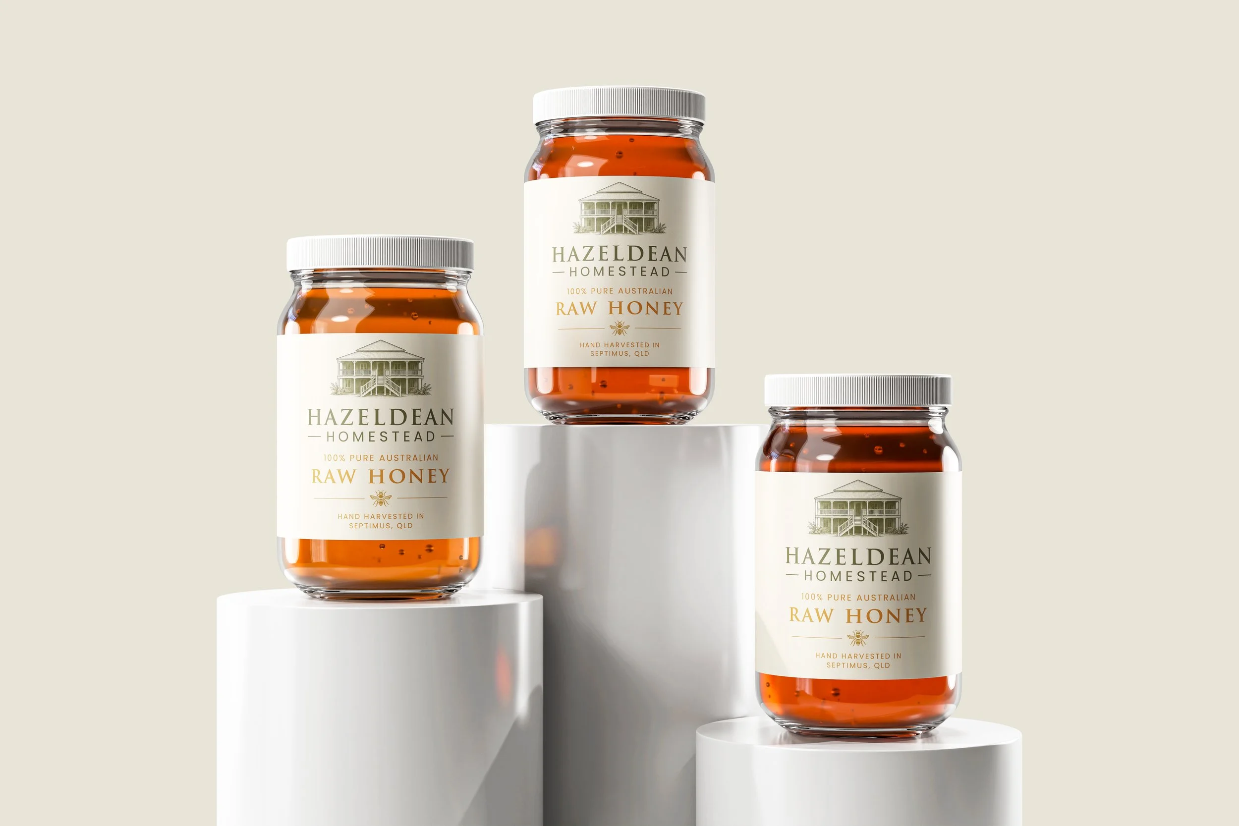 Three jars of Hazeldean Homestead raw honey with white labels featuring an illustration of a house, set on white cylindrical platforms against a beige background.