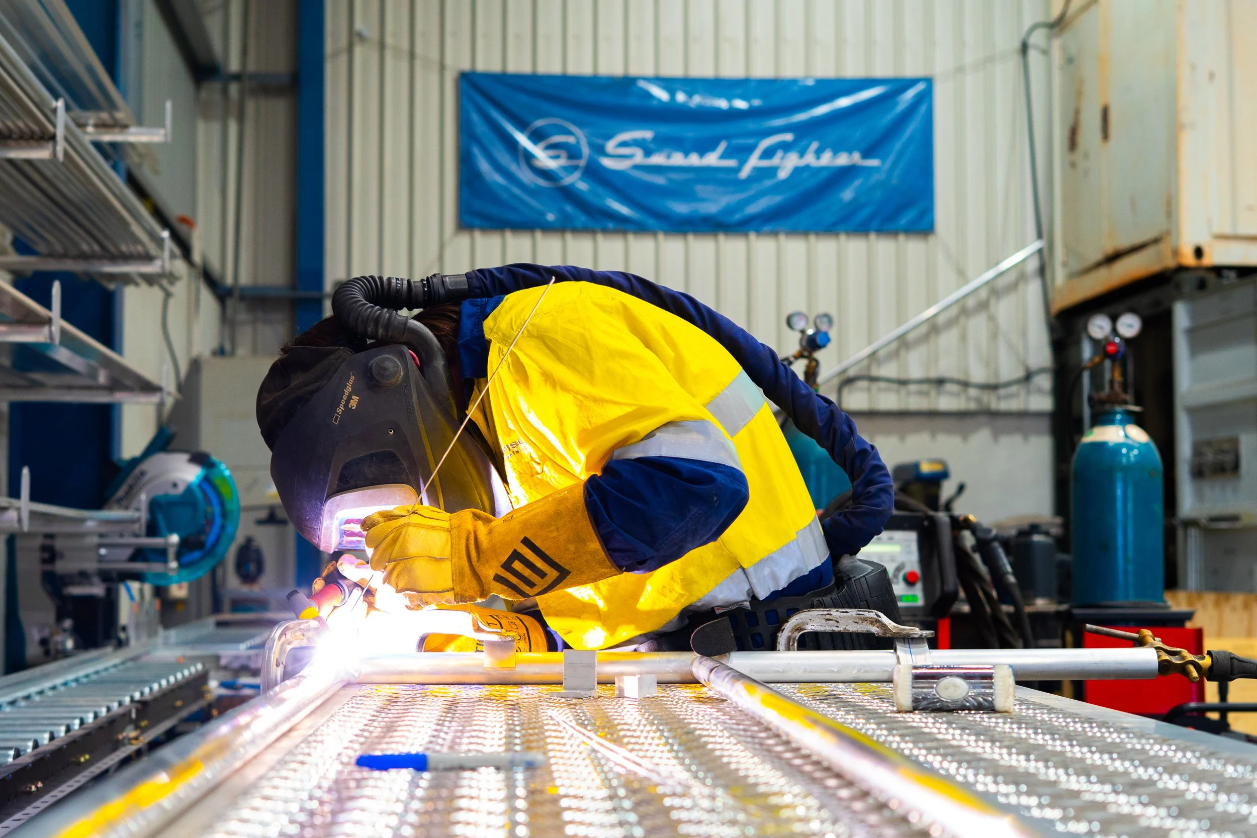 A worker in safety gear welding metal pipes in an industrial workshop, with tools and gauges on the wall and a blue banner in the background.