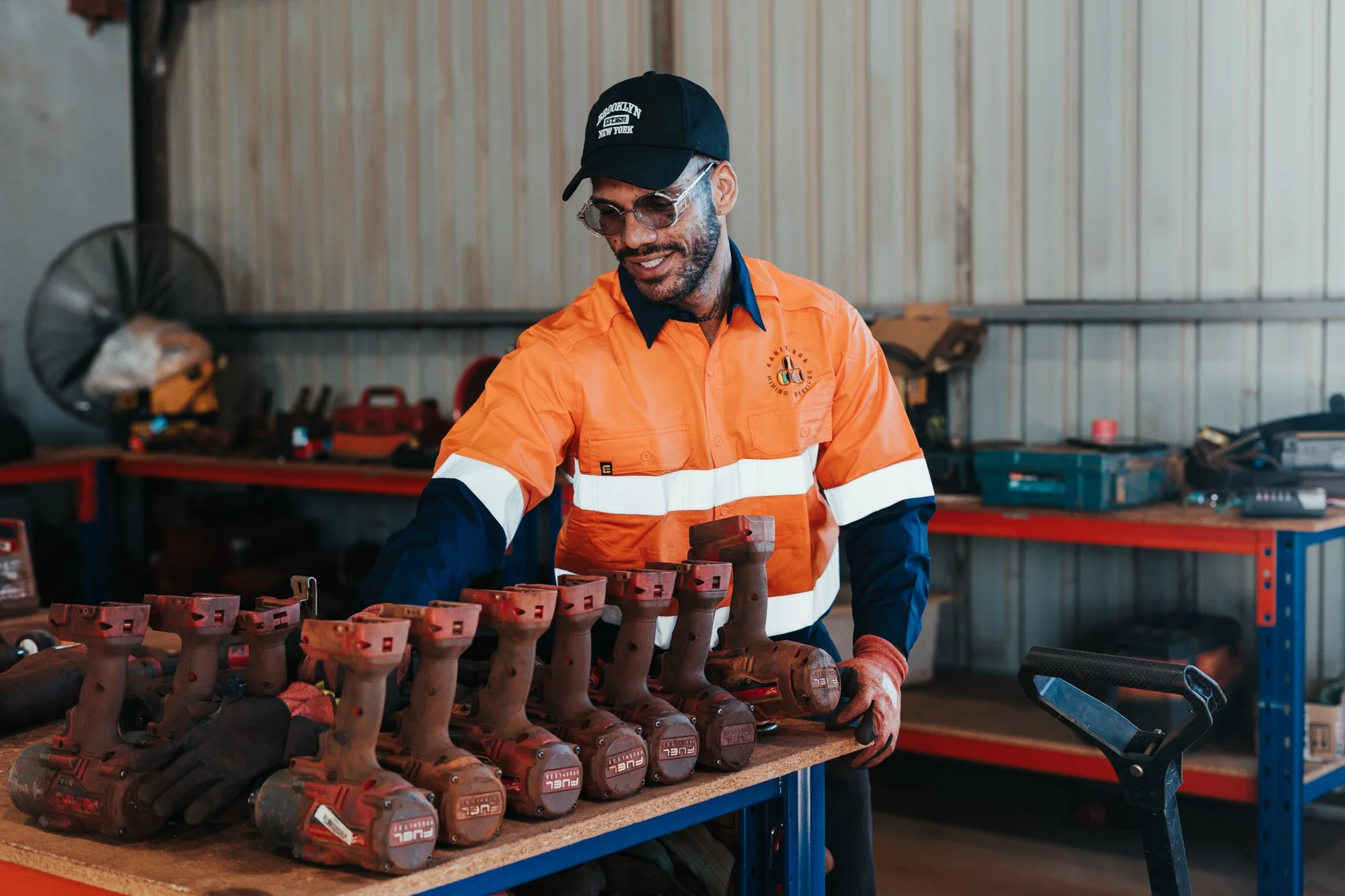 A man in safety glasses and an orange uniform stands in a workshop, organizing work boots on a table.