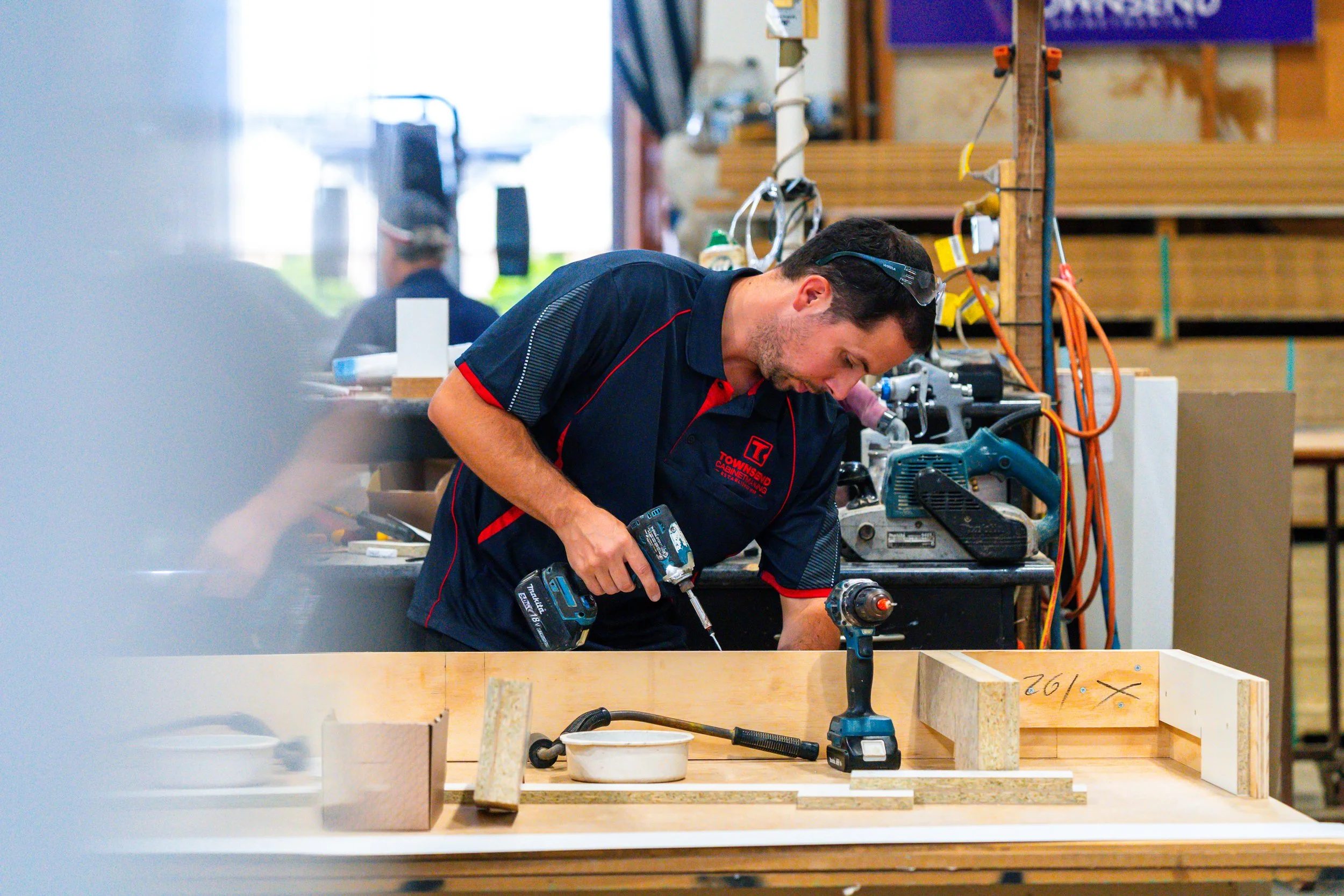 A man working on a woodworking project indoors, using a power drill on a piece of wood, with various tools and equipment around him.