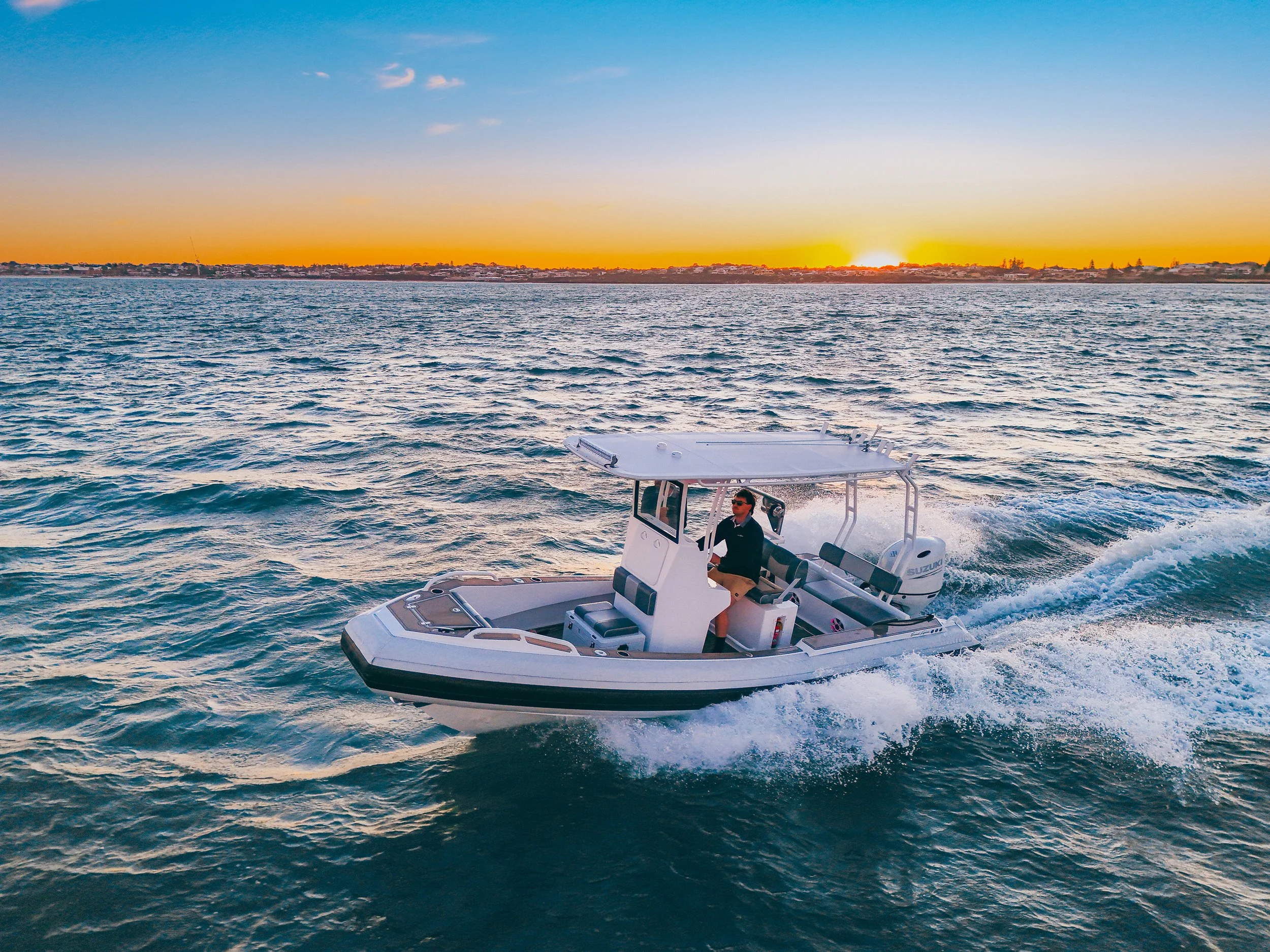 A man driving a small motorboat on the water during sunset, with a cityscape and trees in the background.