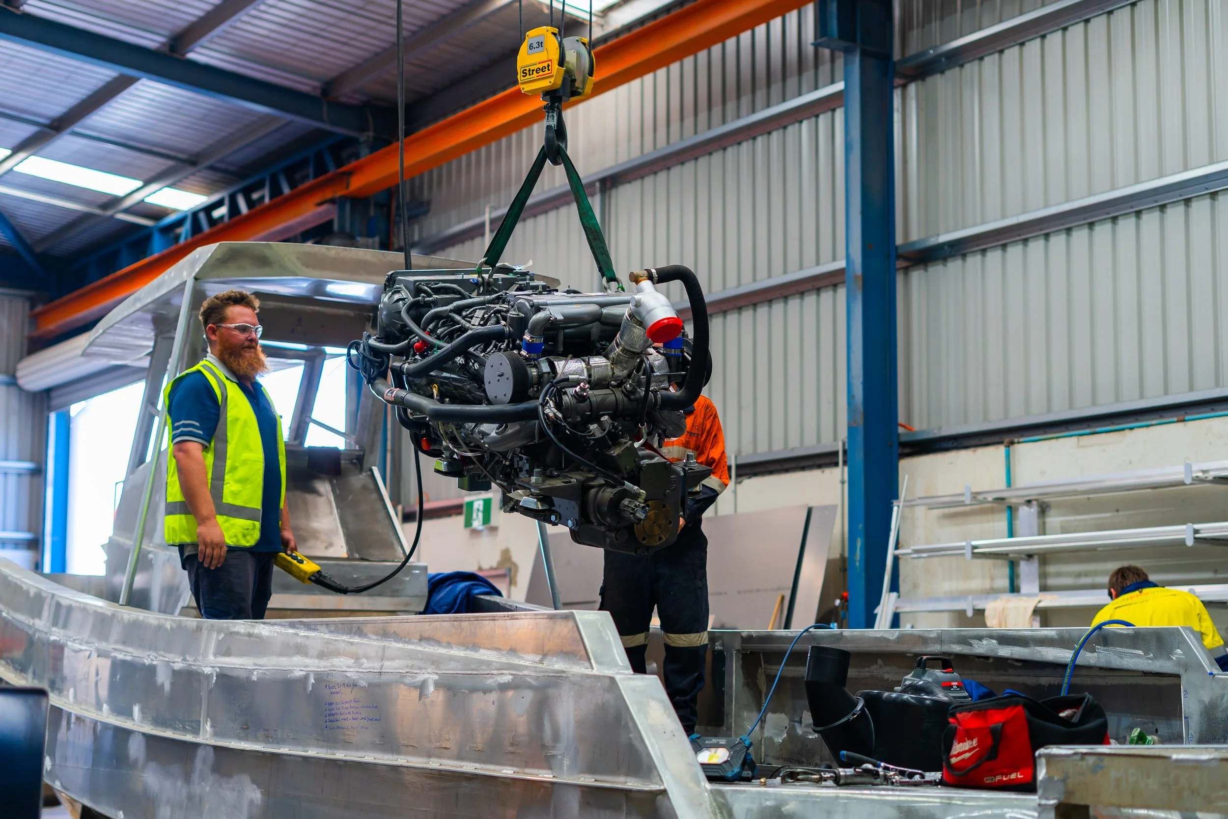 Two men working inside an industrial facility, one of whom is operating a hoist to lift a large engine or machinery part. The man on the left wears safety glasses and a high-visibility vest, while the man on the right is wearing dark clothing. The background includes metal walls, a ceiling with skylights, and industrial equipment.