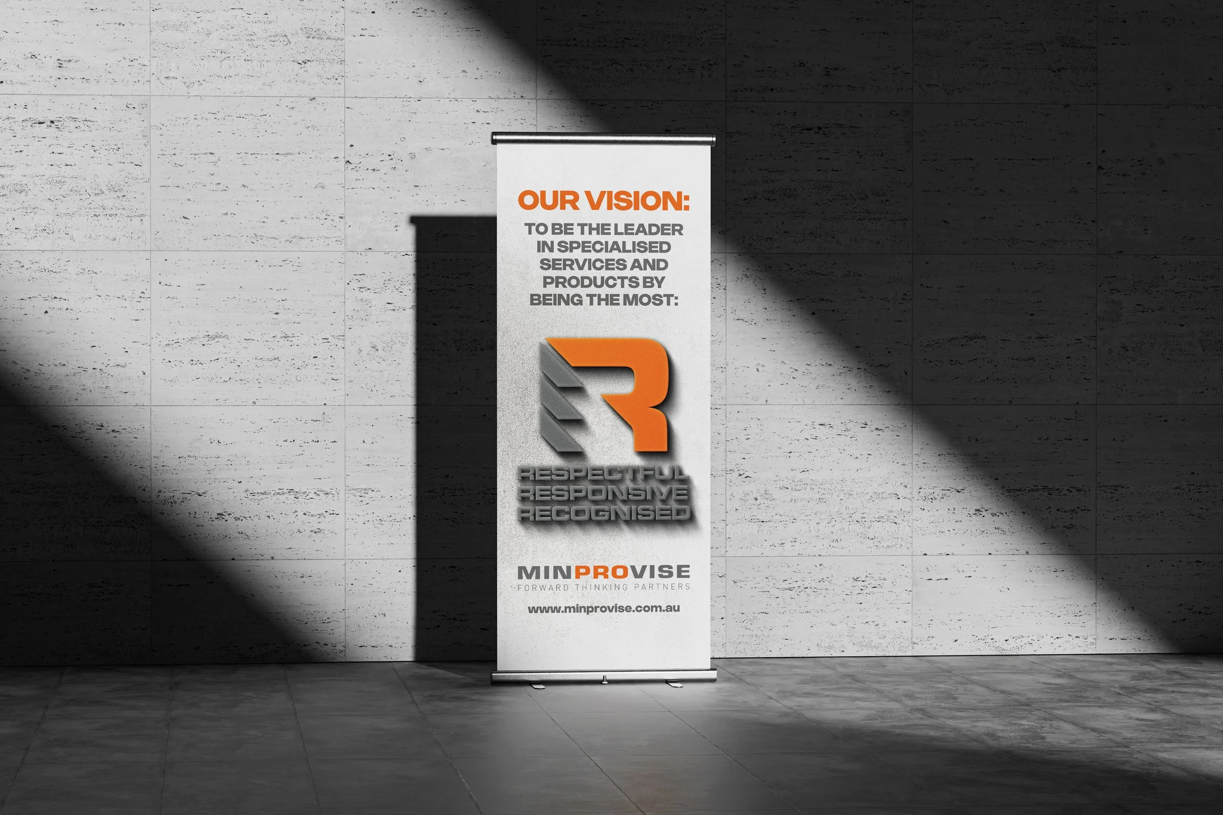 A vertical banner stand with a white background and orange and gray text, placed on a gray tiled floor and wall, displaying a company vision statement and logo.