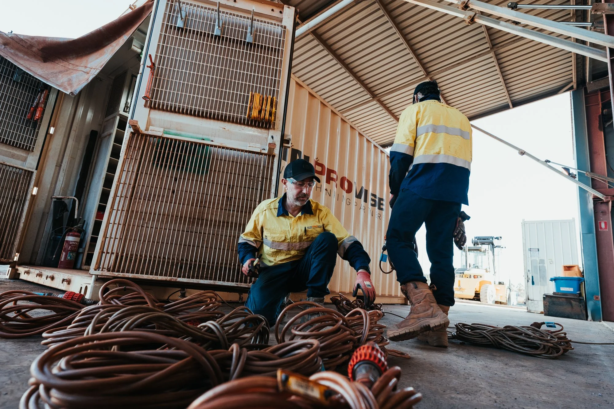 Workers in yellow and navy uniforms wiring or coiling copper cables in a shipping container, with a large industrial container behind them and a forklift in the background.