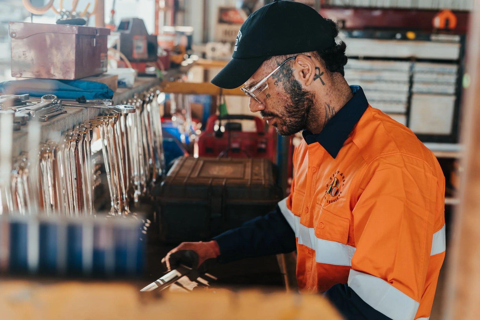 A man wearing glasses, tattoos, and an orange uniform with reflective stripes working in a workshop or garage, surrounded by tools and equipment.