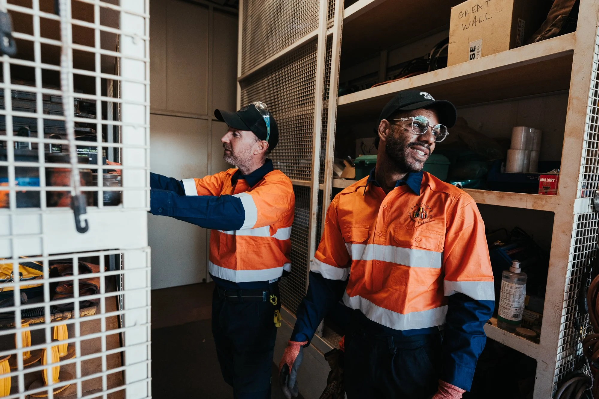 Two workers wearing high-visibility orange and navy uniforms and black caps working in a warehouse or storage room with shelves behind them. One is reaching into a cage, and the other is smiling while looking away.