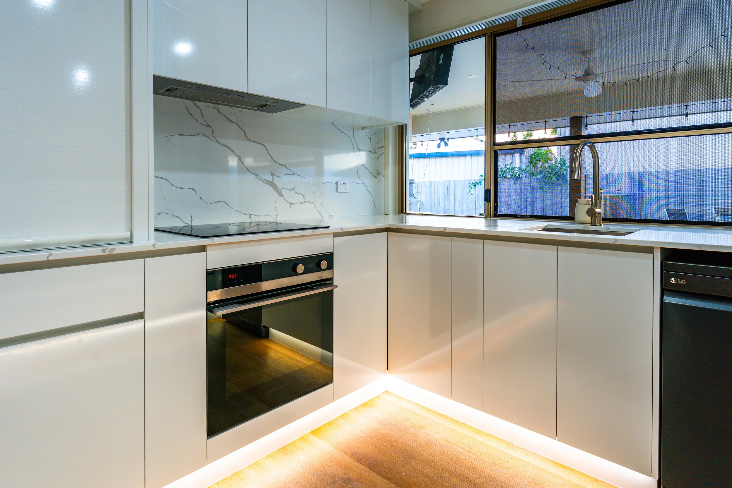 Modern kitchen with white cabinets, marble backsplash, and a large window overlooking an outdoor area with string lights and greenery.