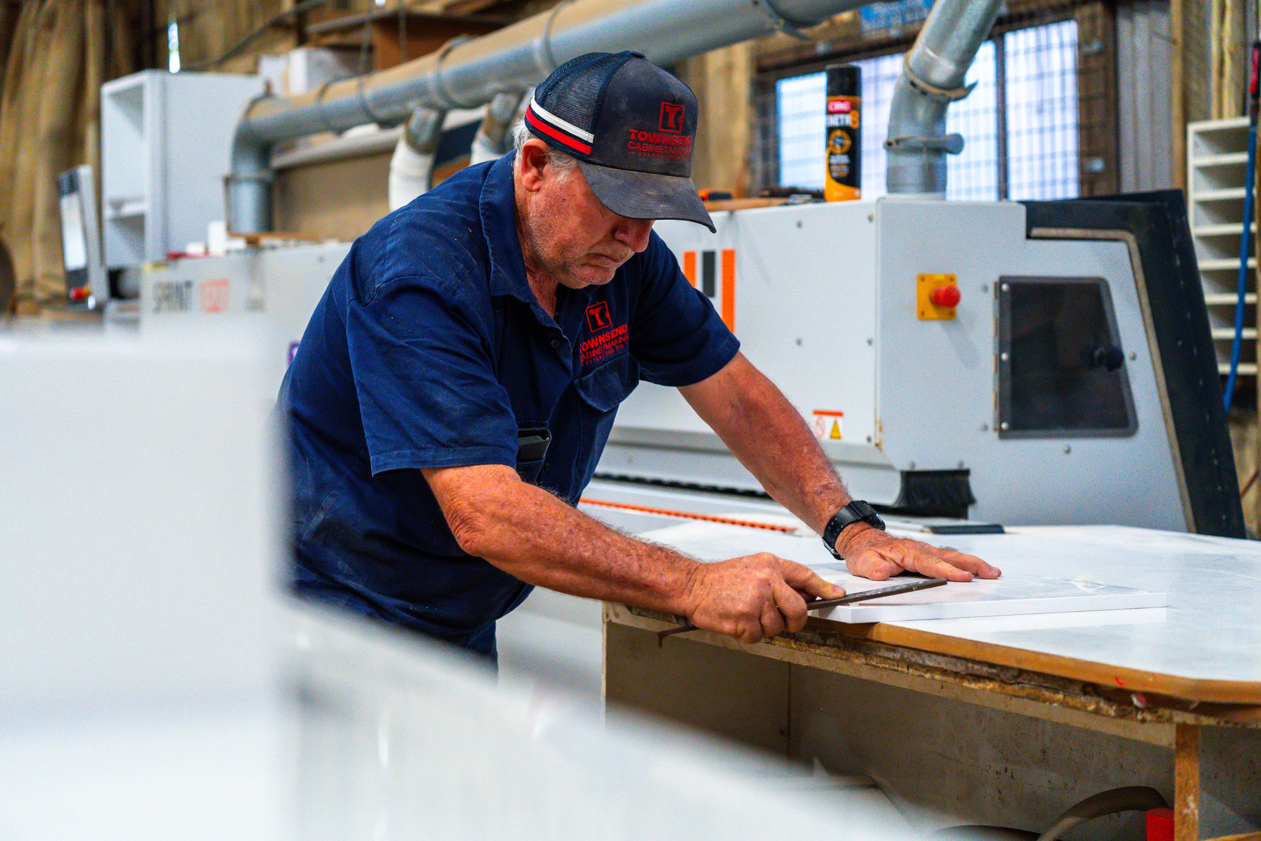 A man working in a woodworking shop, wearing a hat and a blue work shirt, leaning over a workbench with a large woodworking machine in the background.