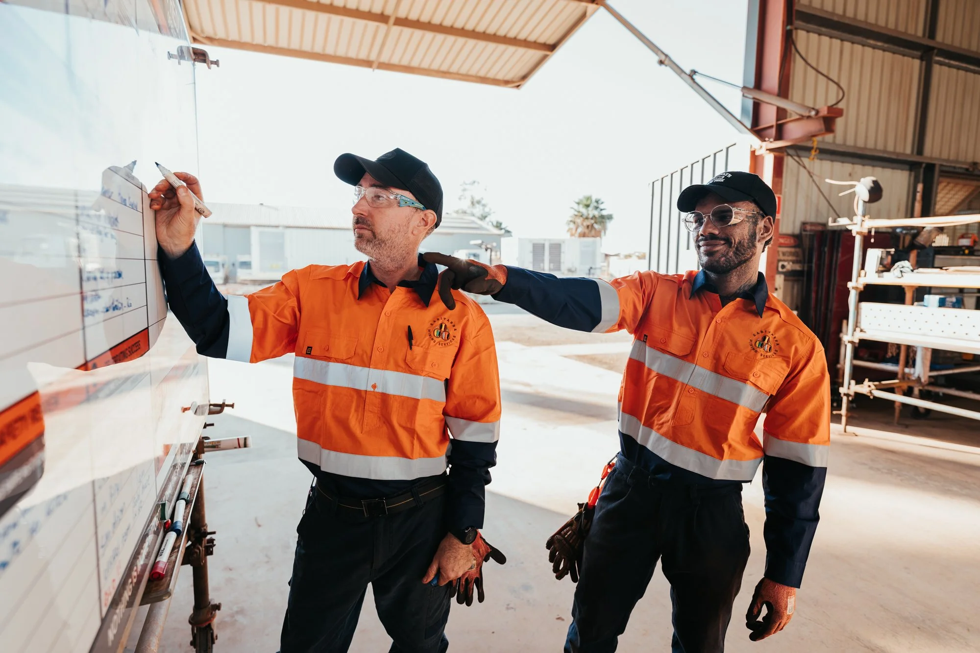 Two male construction workers wearing orange safety vests, gloves, and protective glasses, are working outdoors under a large metal roof. One is writing on a whiteboard with a marker, and the other is standing beside him with a hand on his shoulder, looking at the whiteboard.