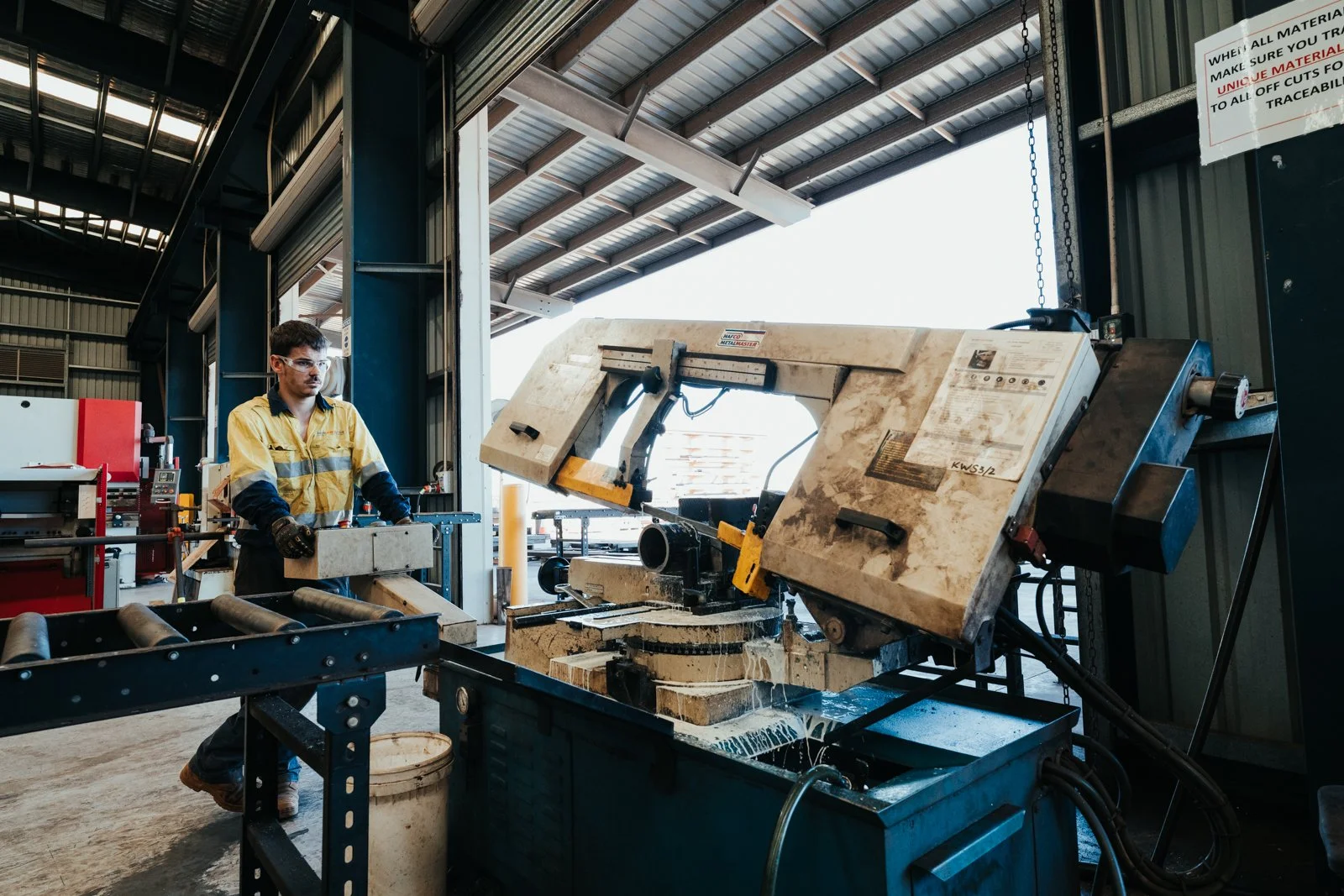 A worker in safety glasses and a yellow and black safety uniform operating a large industrial stone cutting saw inside a warehouse.