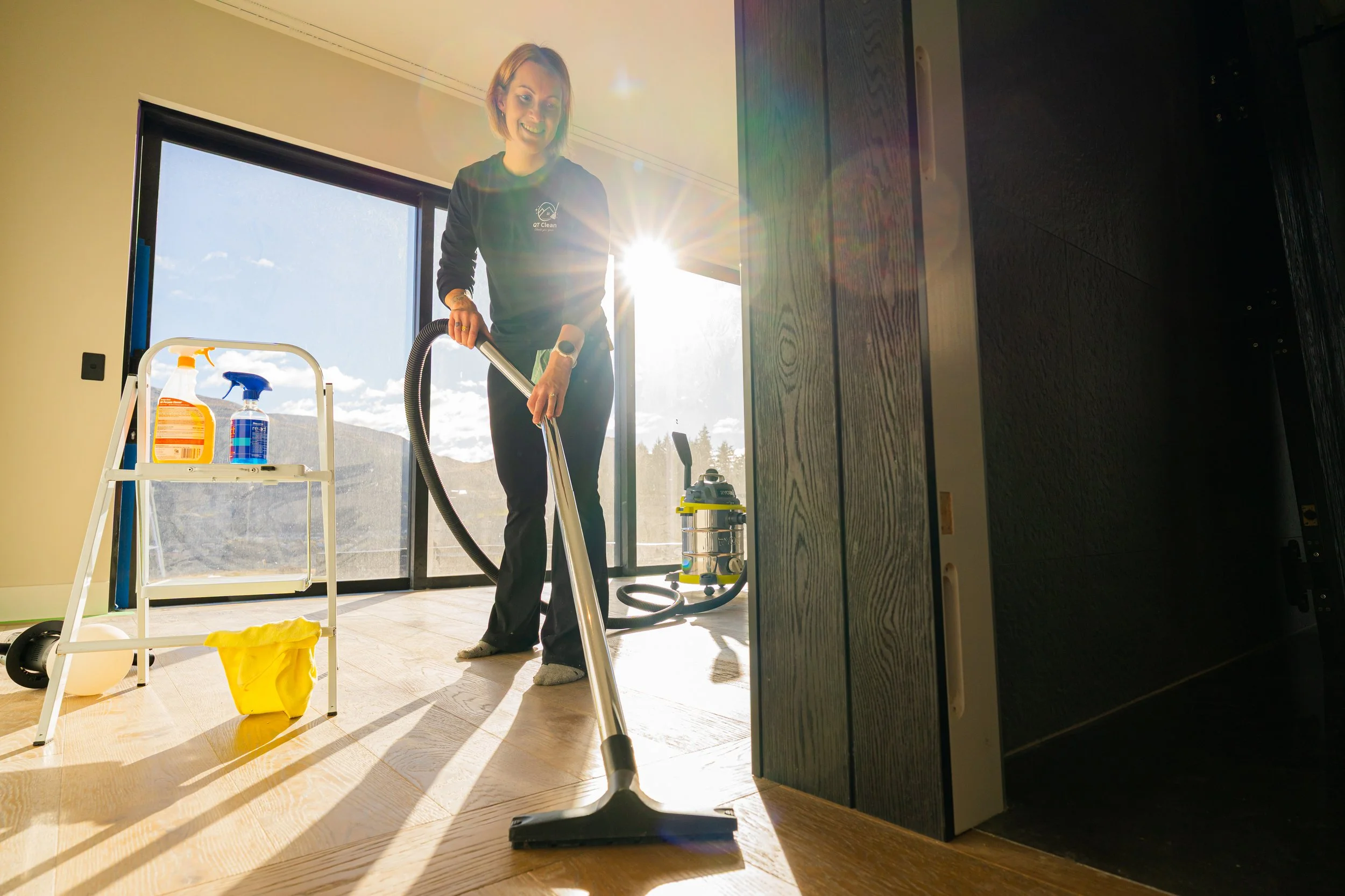 A woman vacuuming a wooden floor in a room with large window, sunlight streaming in, cleaning supplies on a small white cart, and a yellow trash bag on the floor.