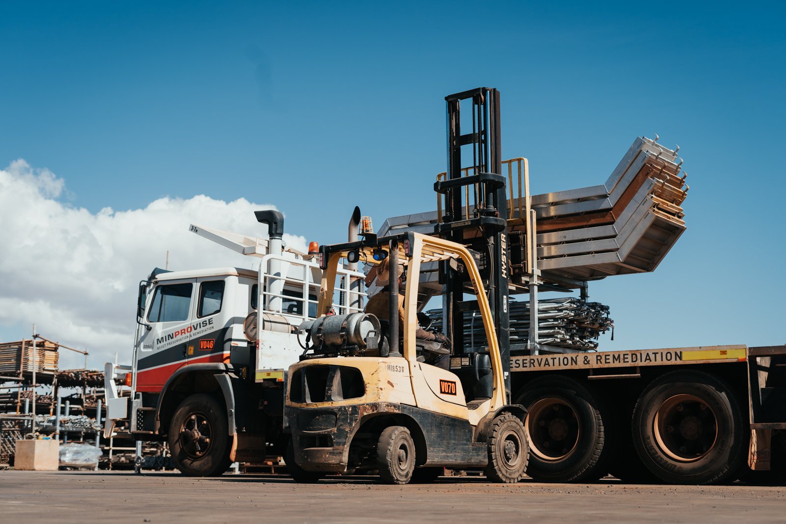 A forklift lifting metal frames on a flatbed truck in an outdoor industrial area under a blue sky.