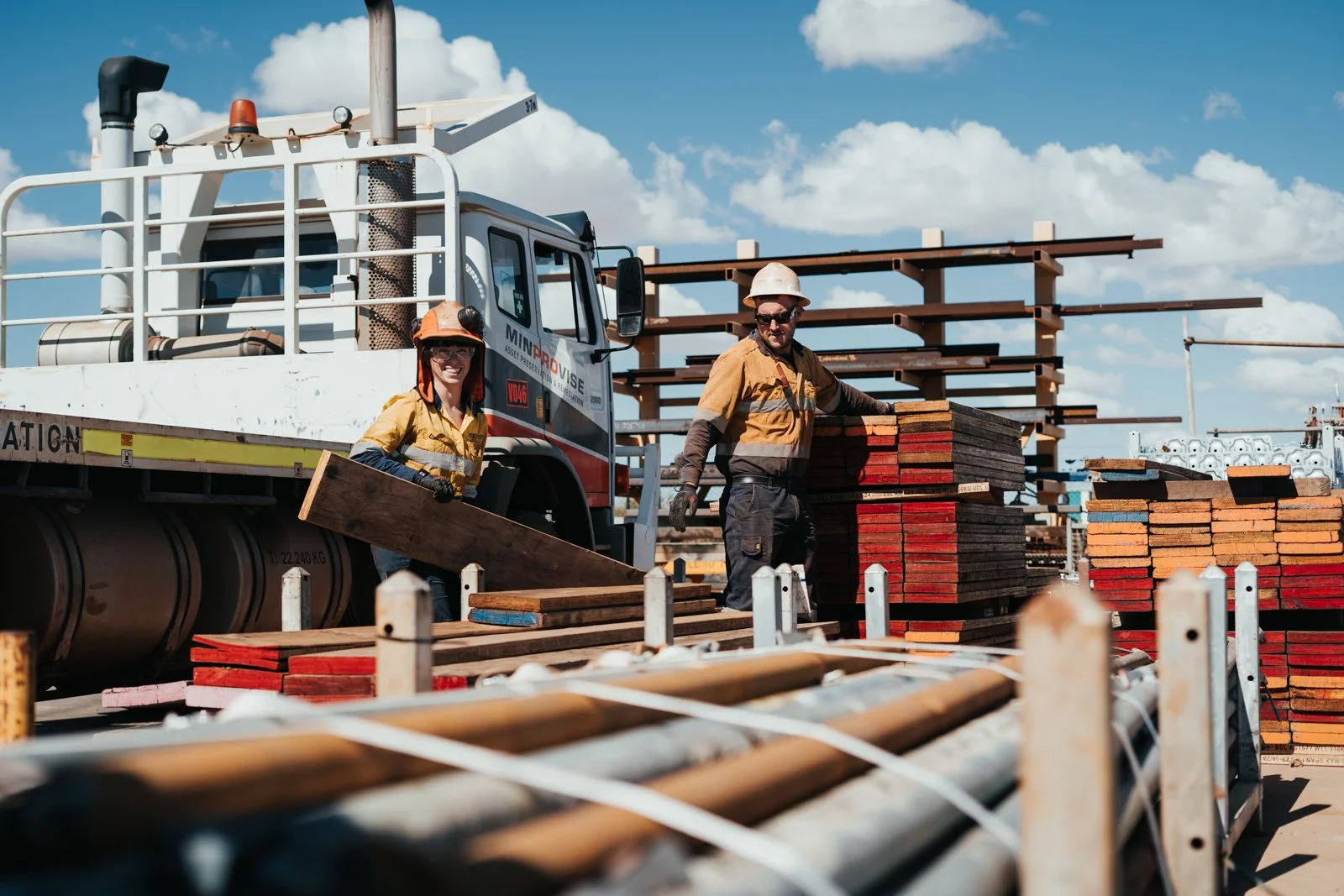 Two construction workers loading wooden planks onto a truck at a construction site under a blue sky with clouds.