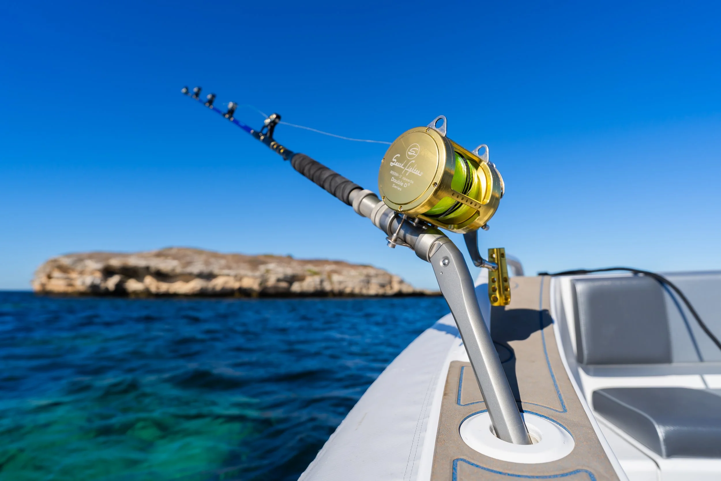 A fishing rod with a gold reel mounted on the side of a boat, extending over clear blue water with a rocky island in the background under a bright blue sky.