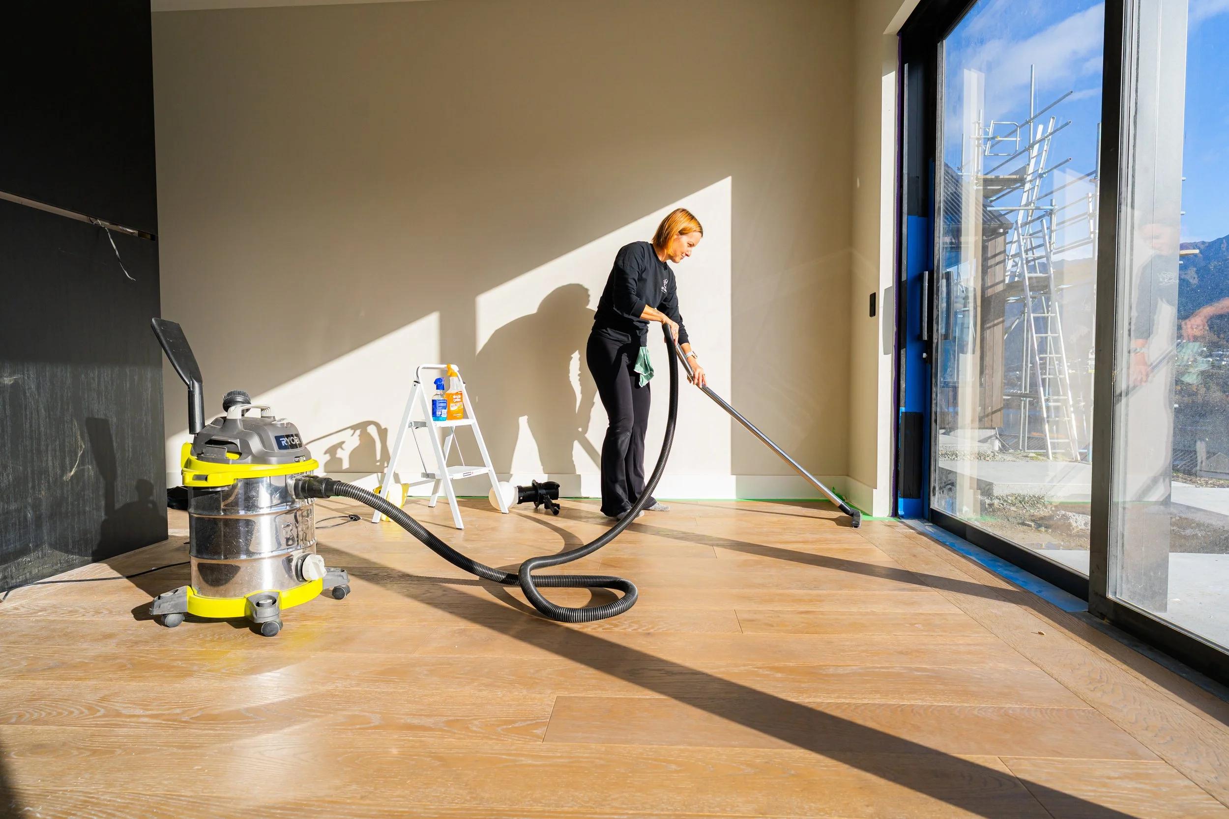 A woman is vacuuming wooden floor inside a room with sunlight coming through large glass doors. She is using a vacuum cleaner, and some cleaning supplies are on a small stepladder nearby.