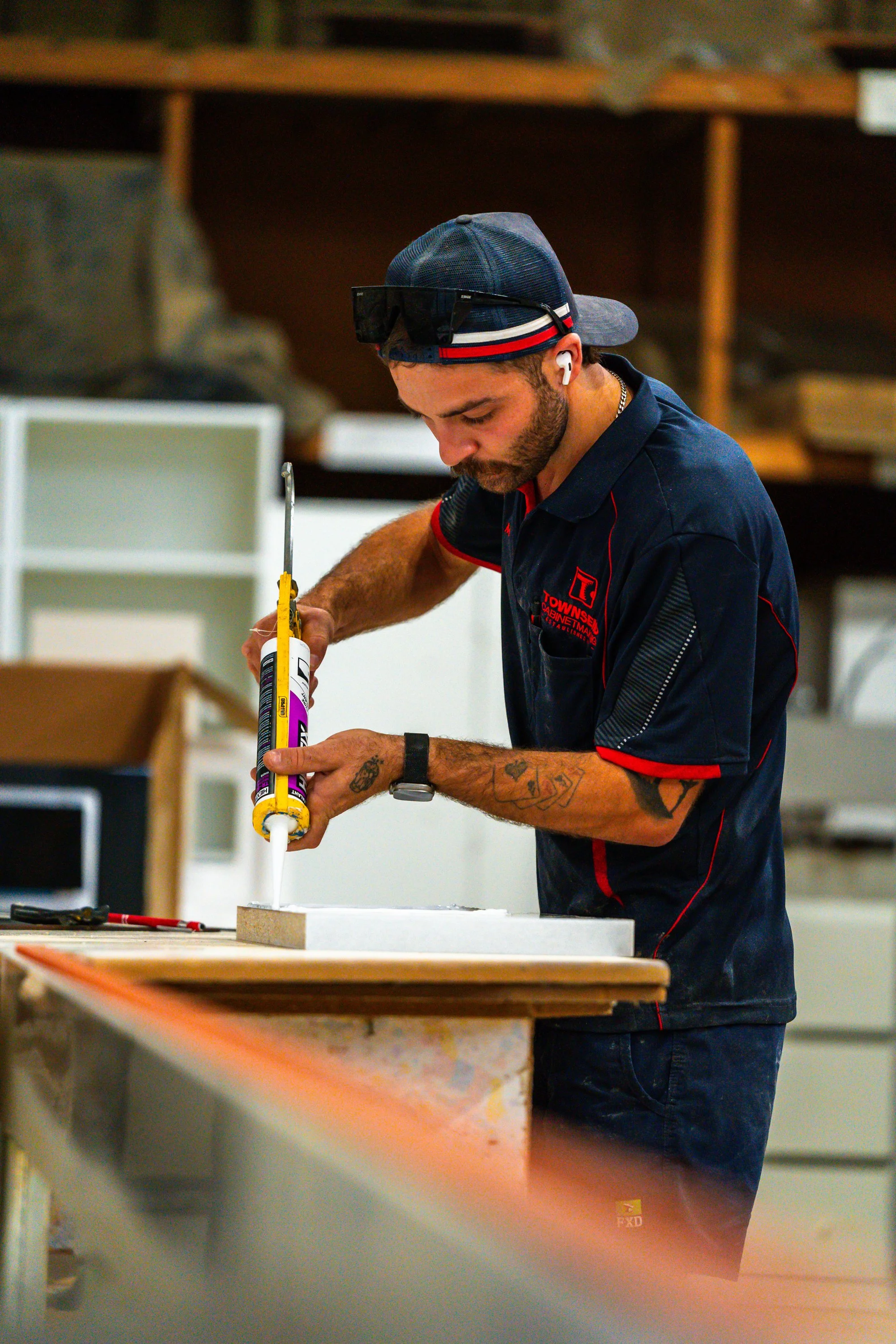A man wearing a cap and sunglasses on his forehead is working at a cluttered workbench. He is applying glue to a piece of material and has tattoos on his forearms.
