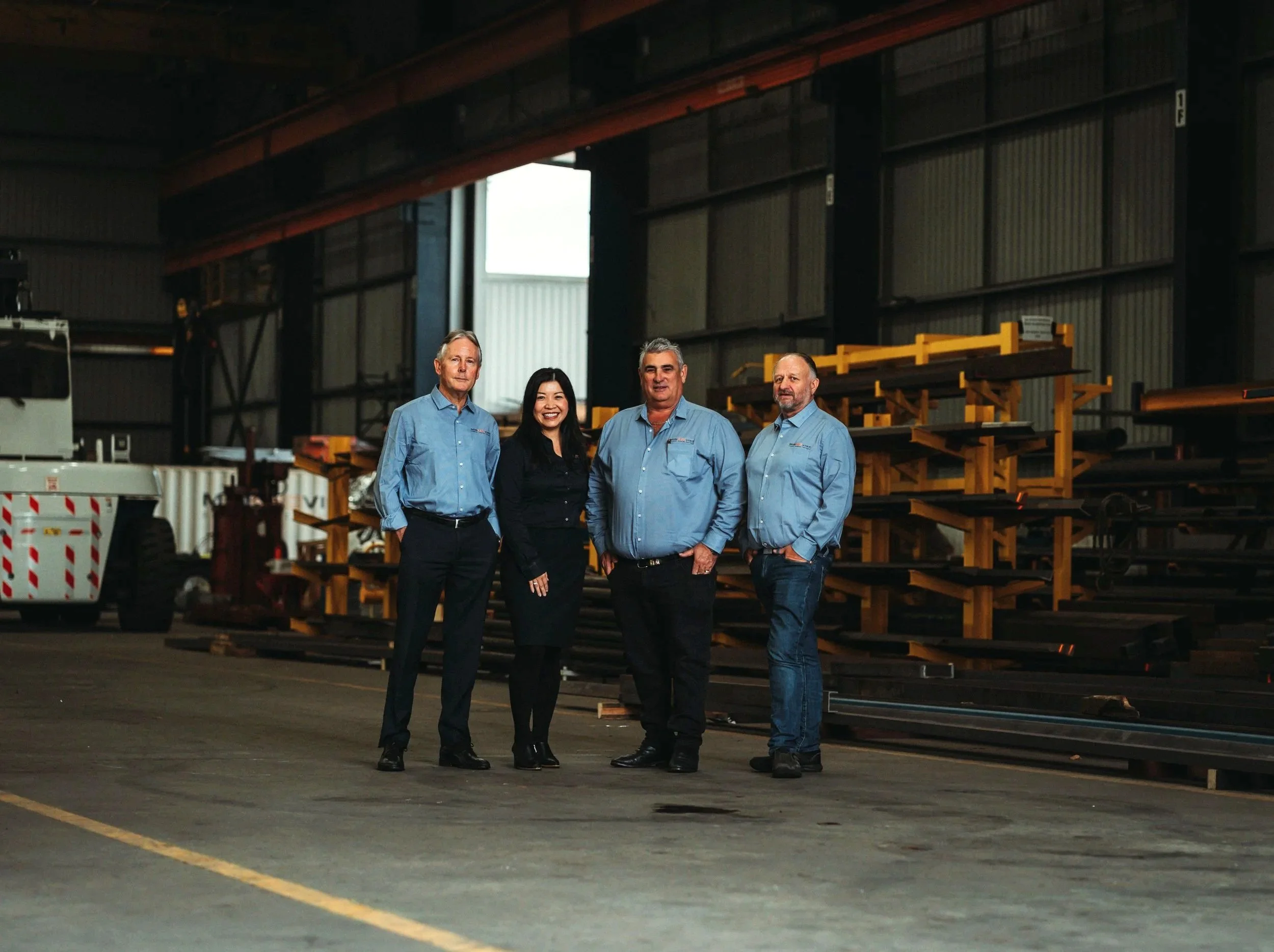 Group of four people standing inside a large industrial warehouse, with metal shelves and equipment in the background.