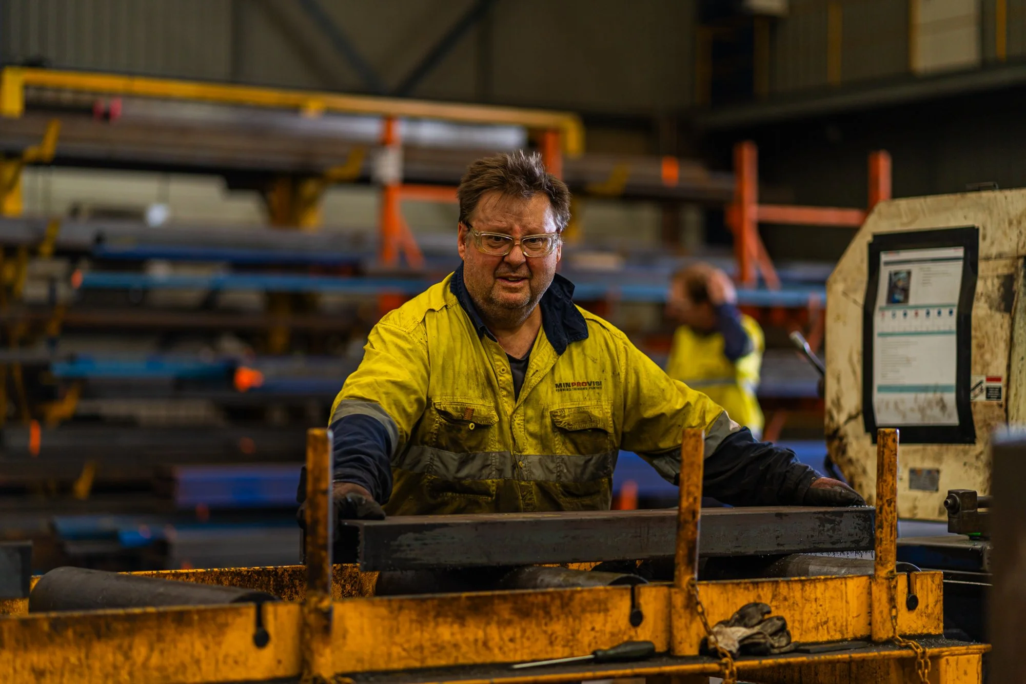 Man wearing safety glasses and yellow work jacket operating heavy machinery in a warehouse setting with metal racks and workers in the background.