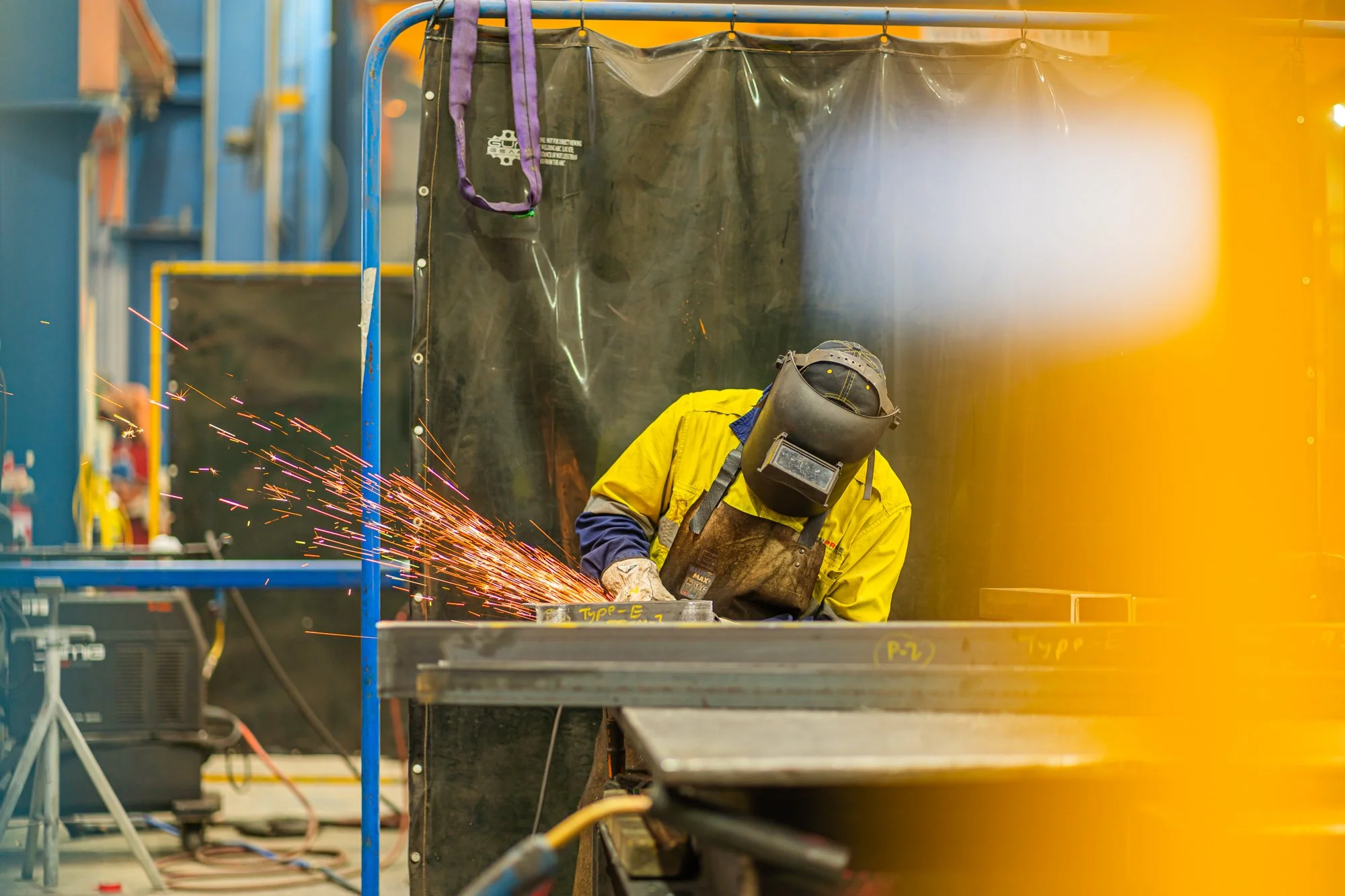 A welder wearing a yellow protective jacket, helmet, and gloves is welding metal on a workbench inside a factory or workshop. Sparks are flying from the welding process, and there are safety barriers and equipment in the background.