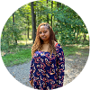 A black woman with long brown sisterlocks standing outdoors in a park with trees and grass, wearing a floral dress.