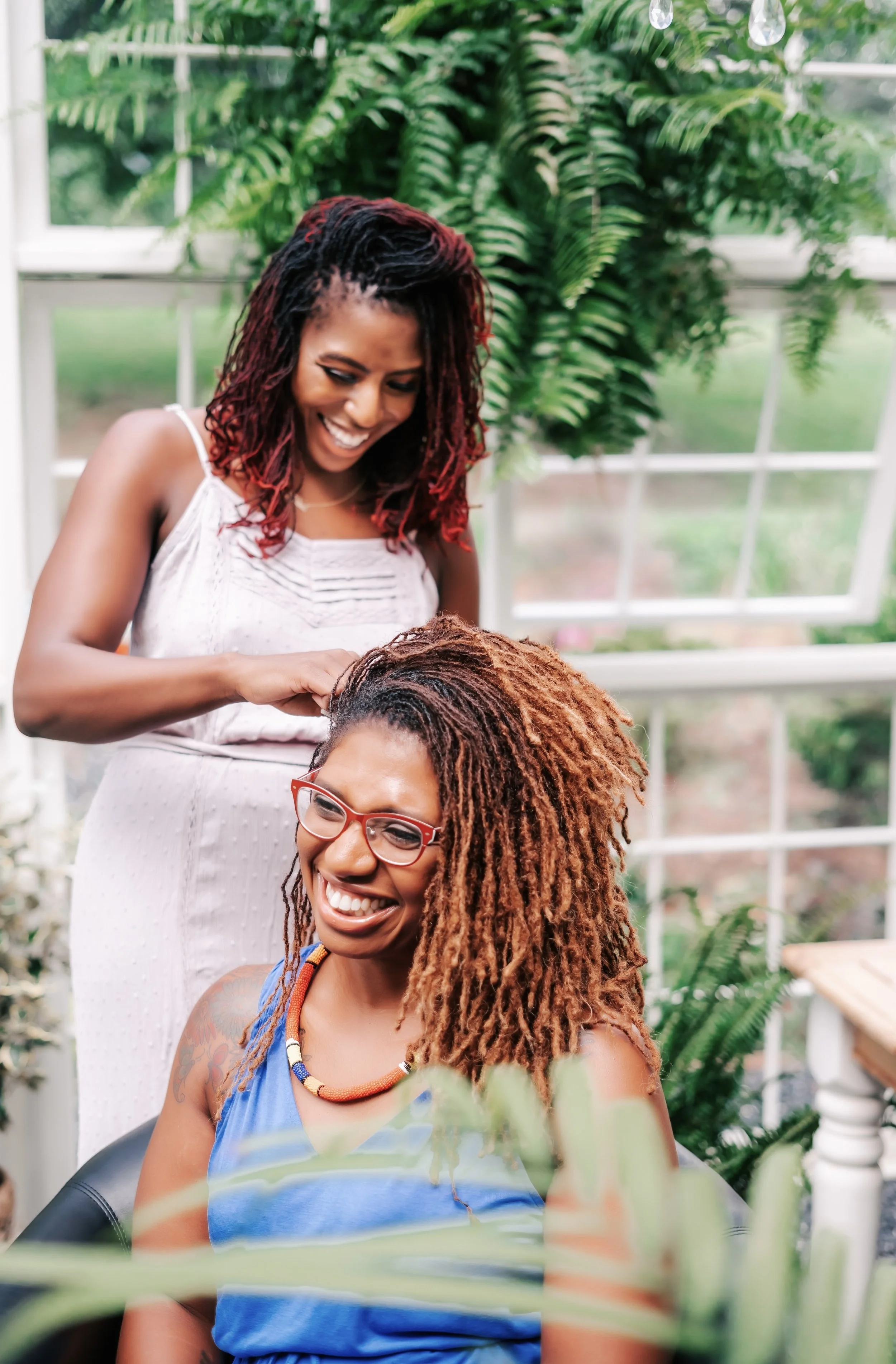 A SISTERLOCKS consultant maintains her clients beautiful long sisterlocks women with sisterlocks while smiling inside a greenhouse filled with green plants and large windows.